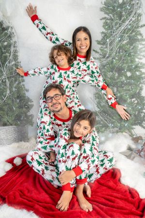 Family wearing matching Christmas tree pajamas pose playfully on red blanket with snowy backdrop.