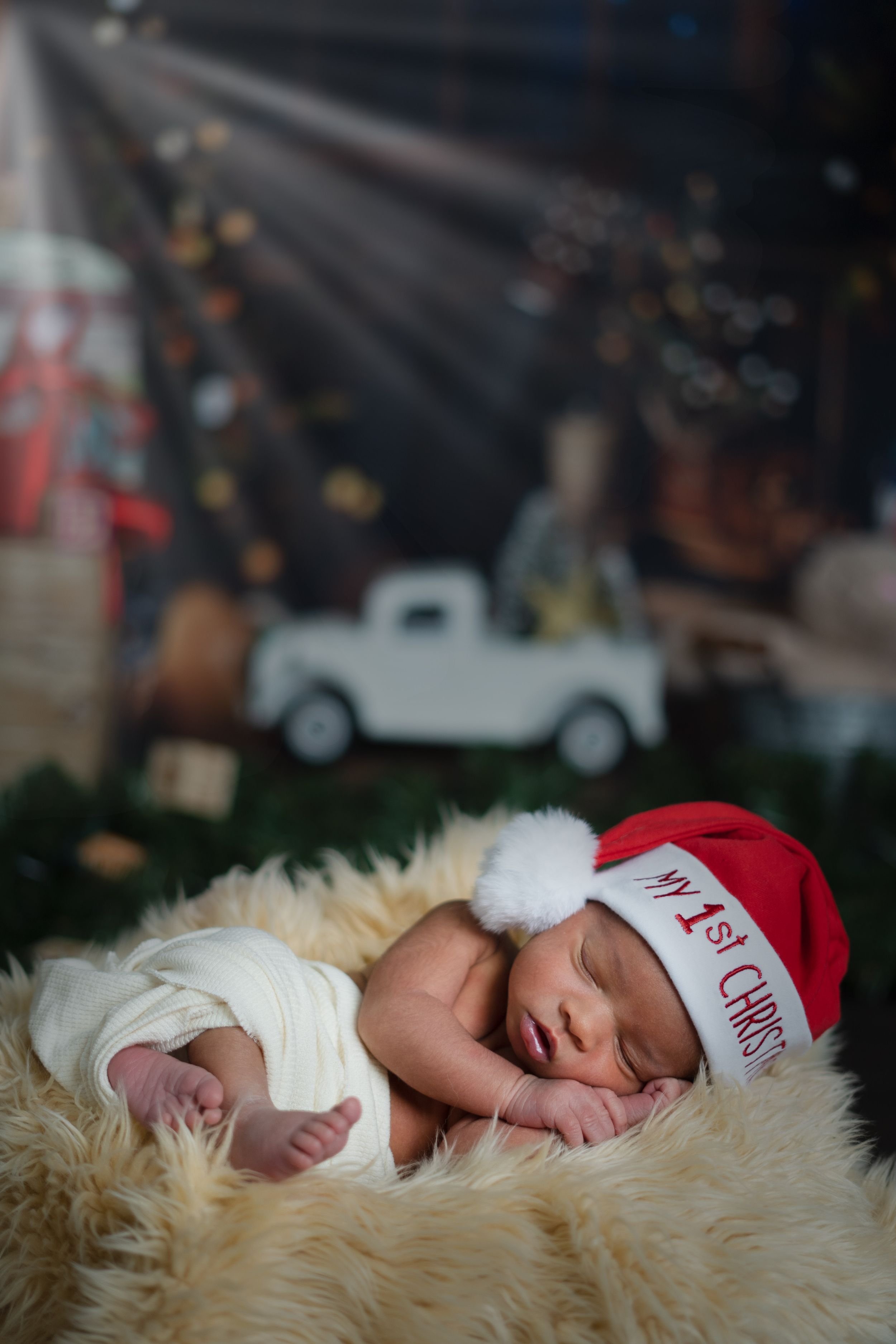 Sleeping baby wearing red Santa hat rests on fluffy cream blanket with vintage decor background.