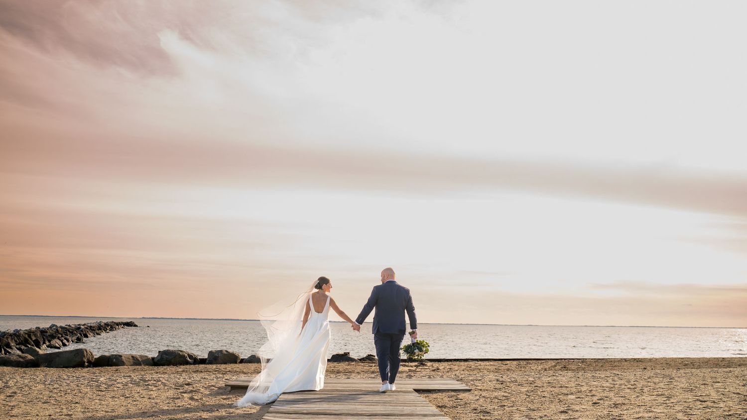 A wedding couple walking on a rocky beach at sunset, holding hands against a pink and orange sky at Land's End