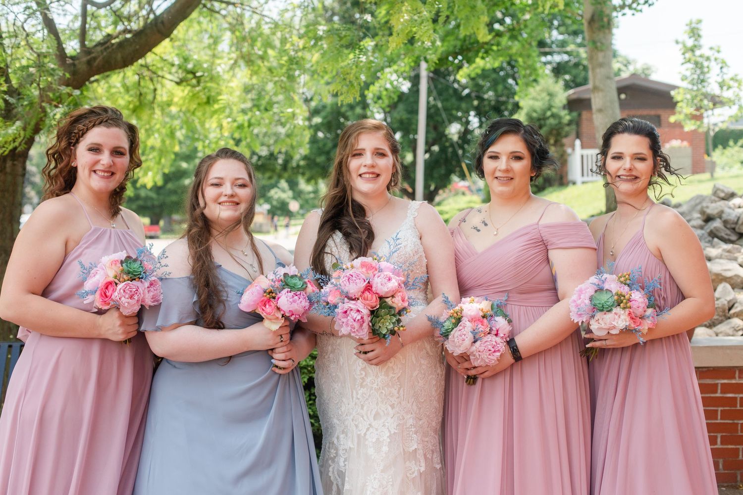 Bridal party in pink and gray dresses holding pink and white bouquets pose outdoors.