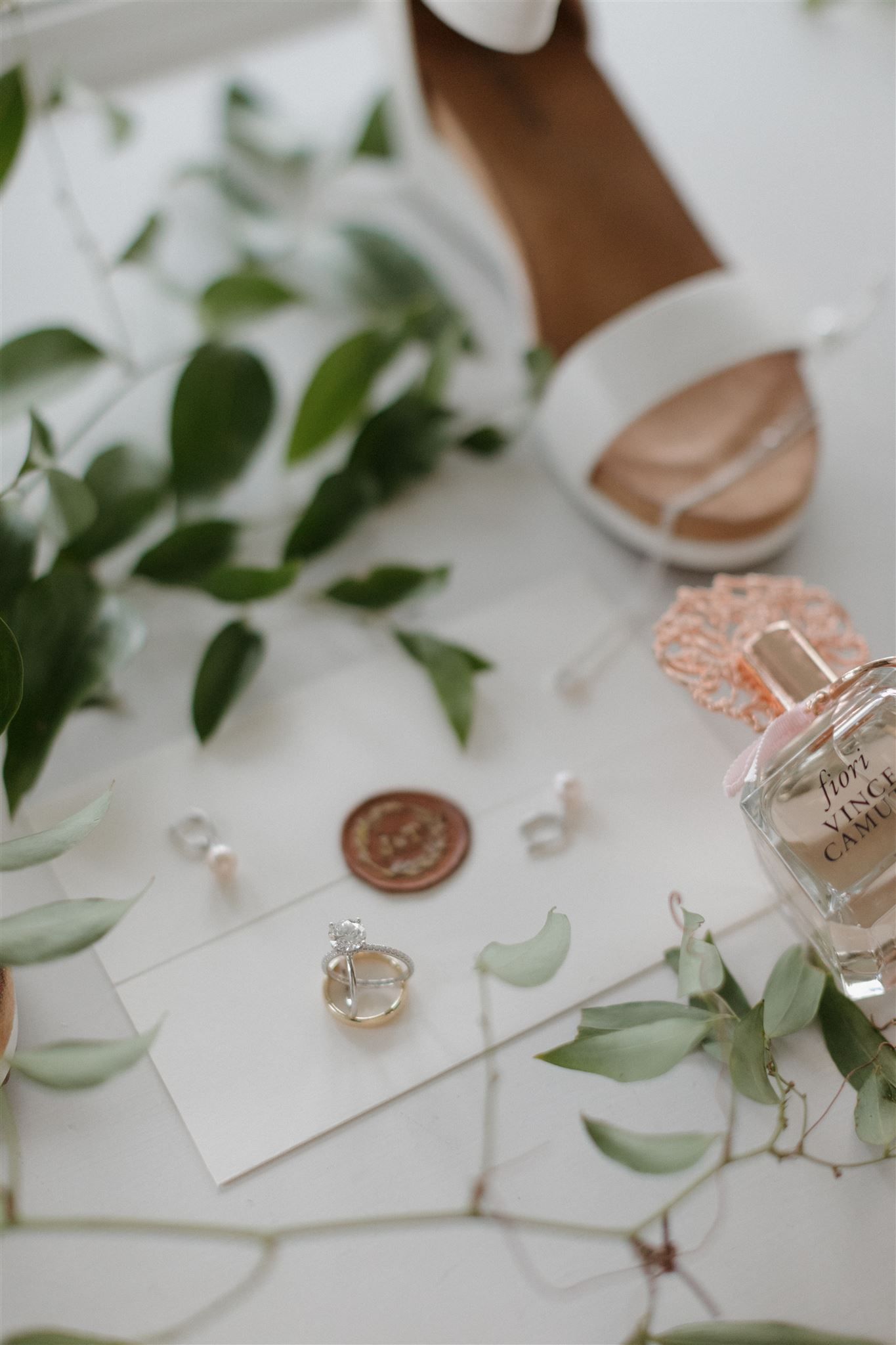 Wedding details including white shoes, delicate greenery, jewelry and a wax seal on a light background.
