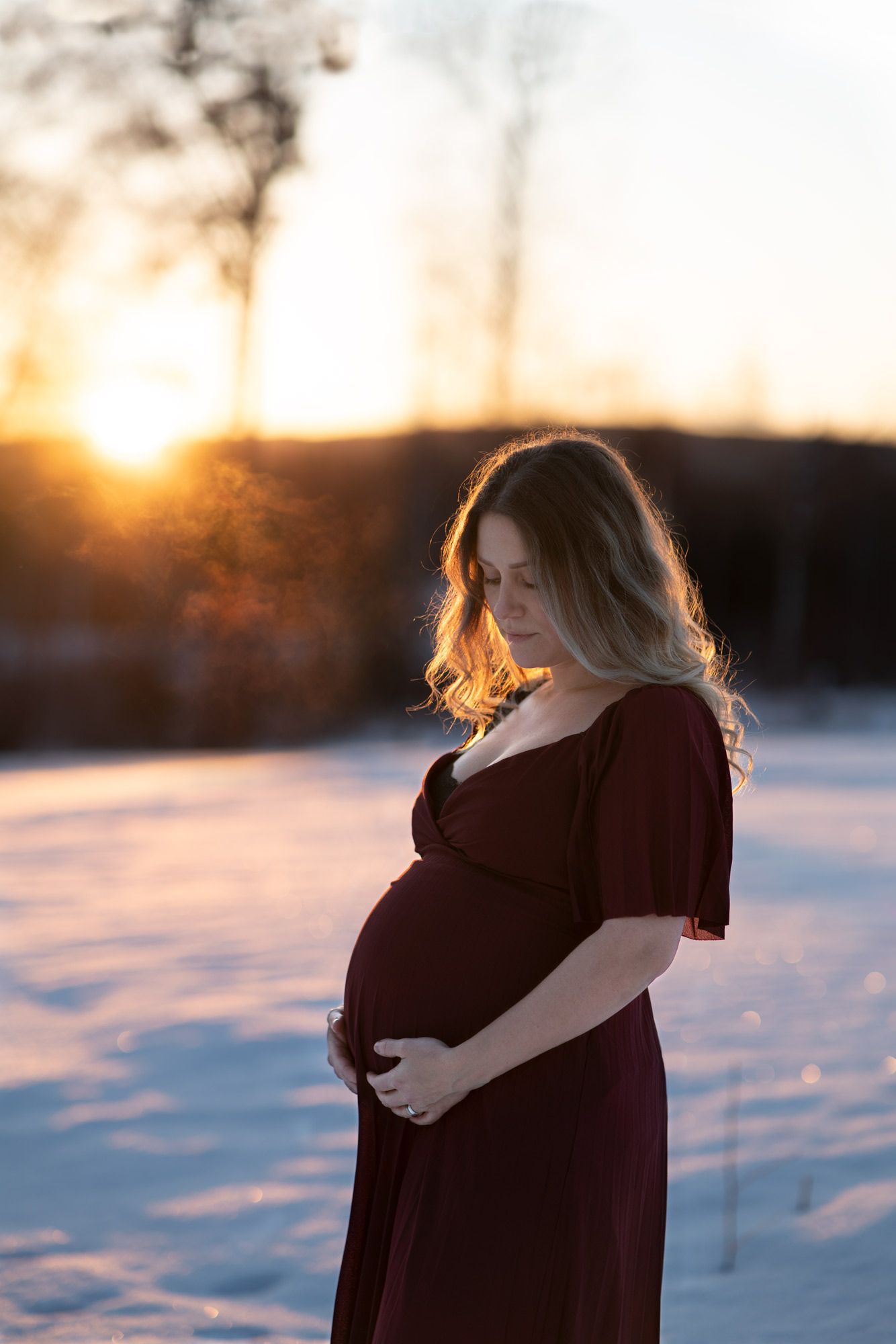 Gravid kvinna i röd klänning står ute i snön i soluppgången