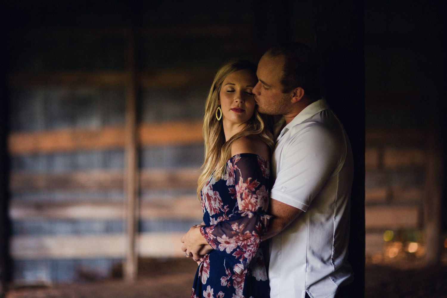 Romantic couple embracing in moody barn setting with wooden wall backdrop.