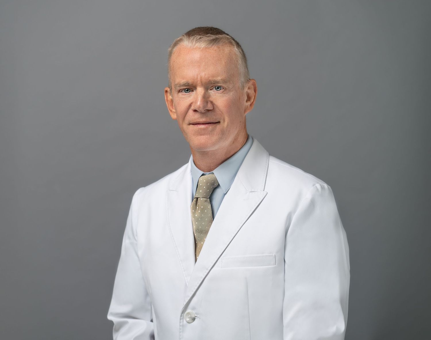 Professional portrait of person in white medical coat and tie against grey background.