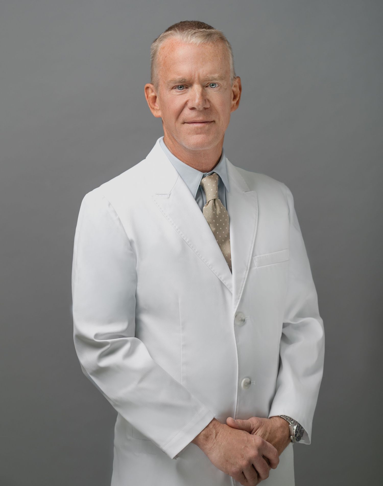 Professional portrait of a medical practitioner wearing a white coat and tie against a gray background.