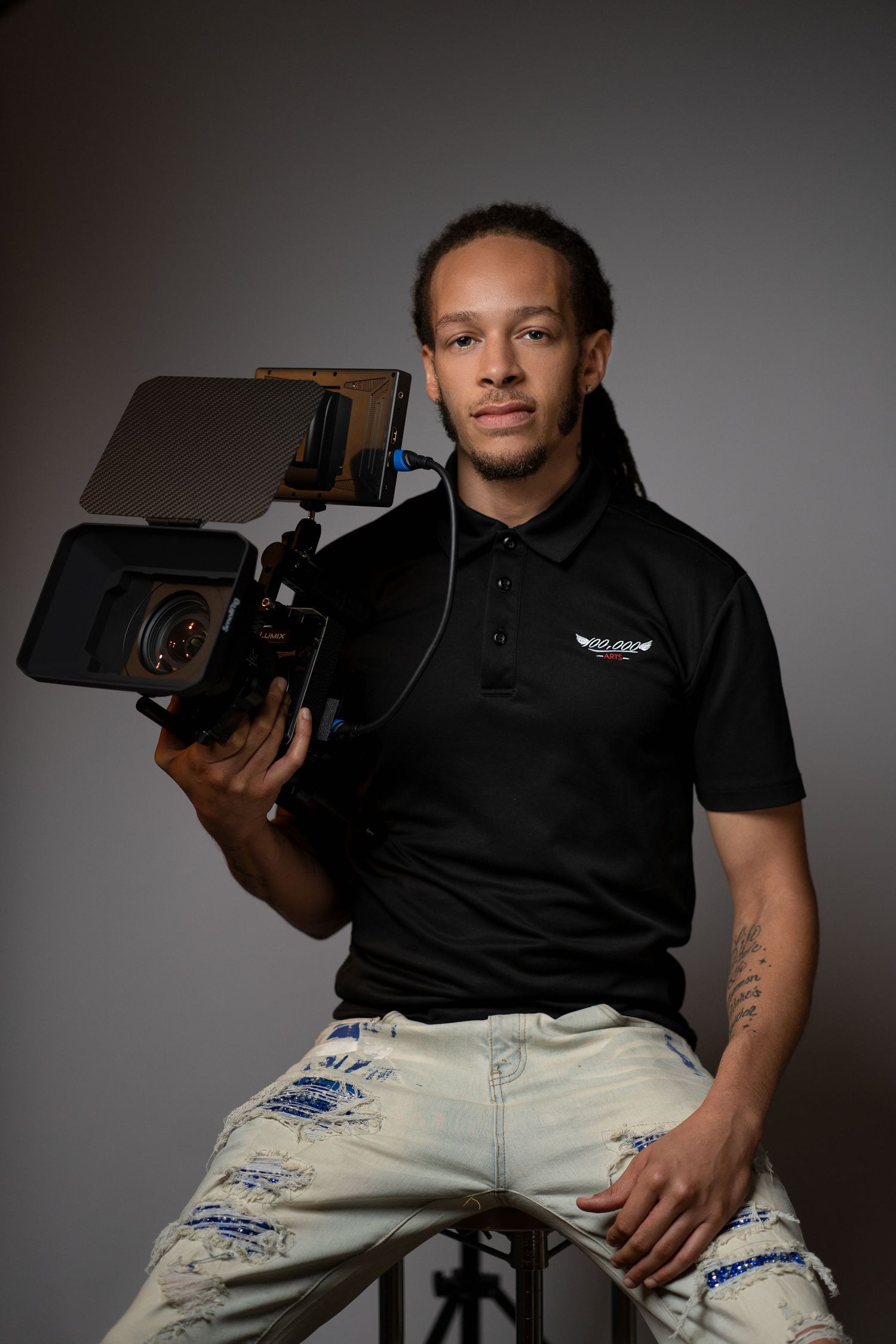 Videographer wearing black polo shirt holding professional camera equipment in studio setting.