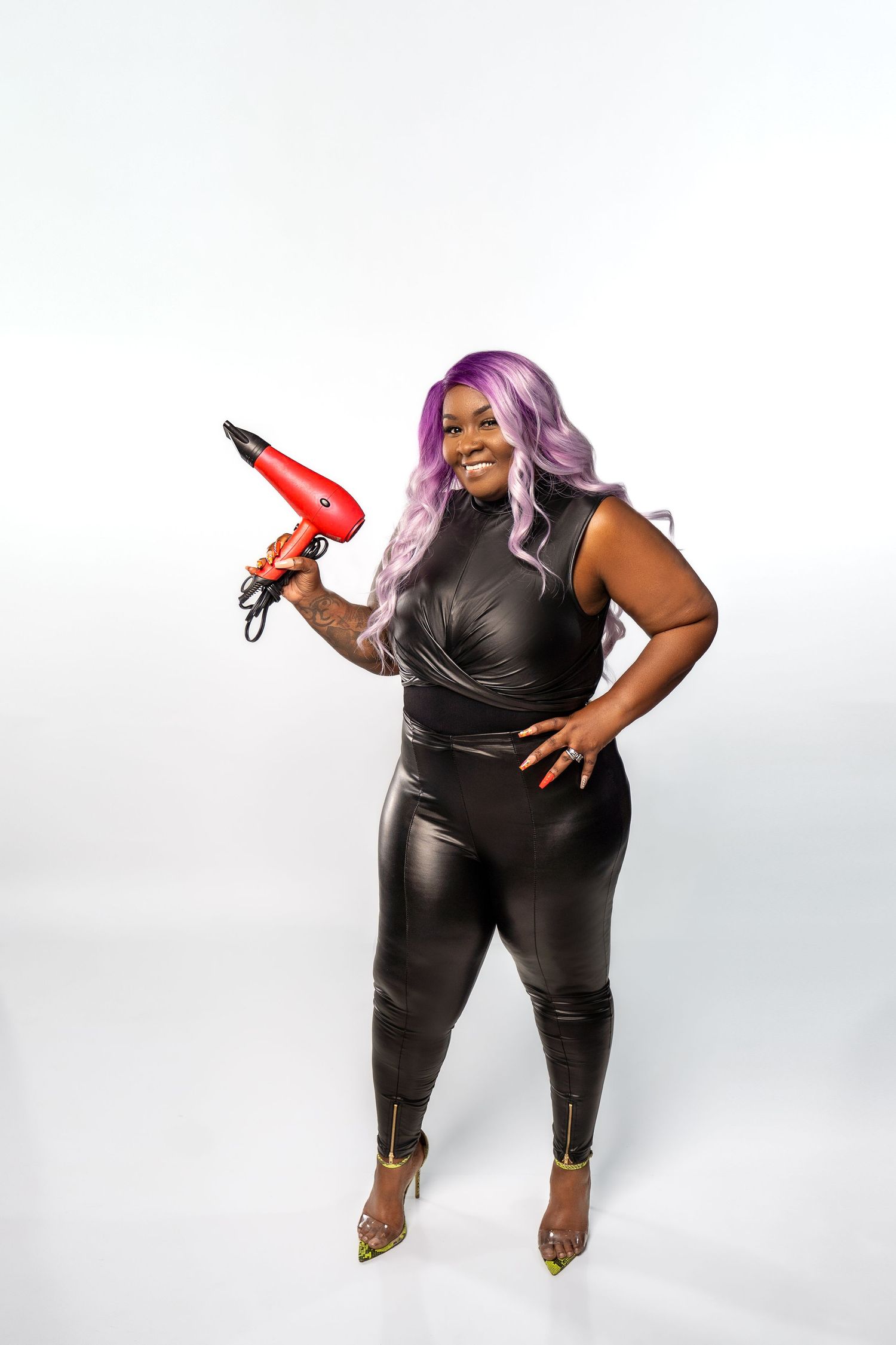 Person with purple hair in black leather outfit holds hair dryer against white background.