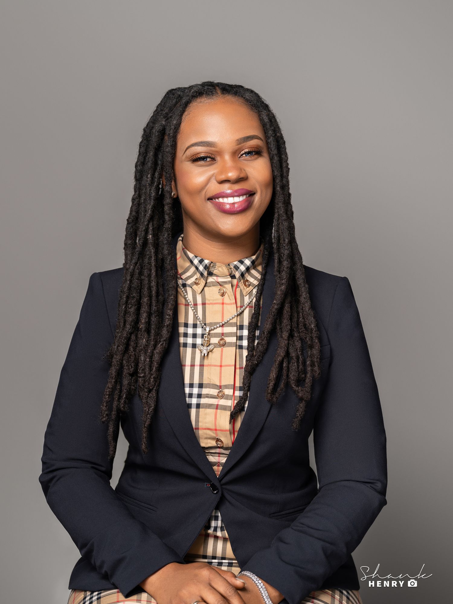 Confident woman in business attire with locs poses against grey studio backdrop.