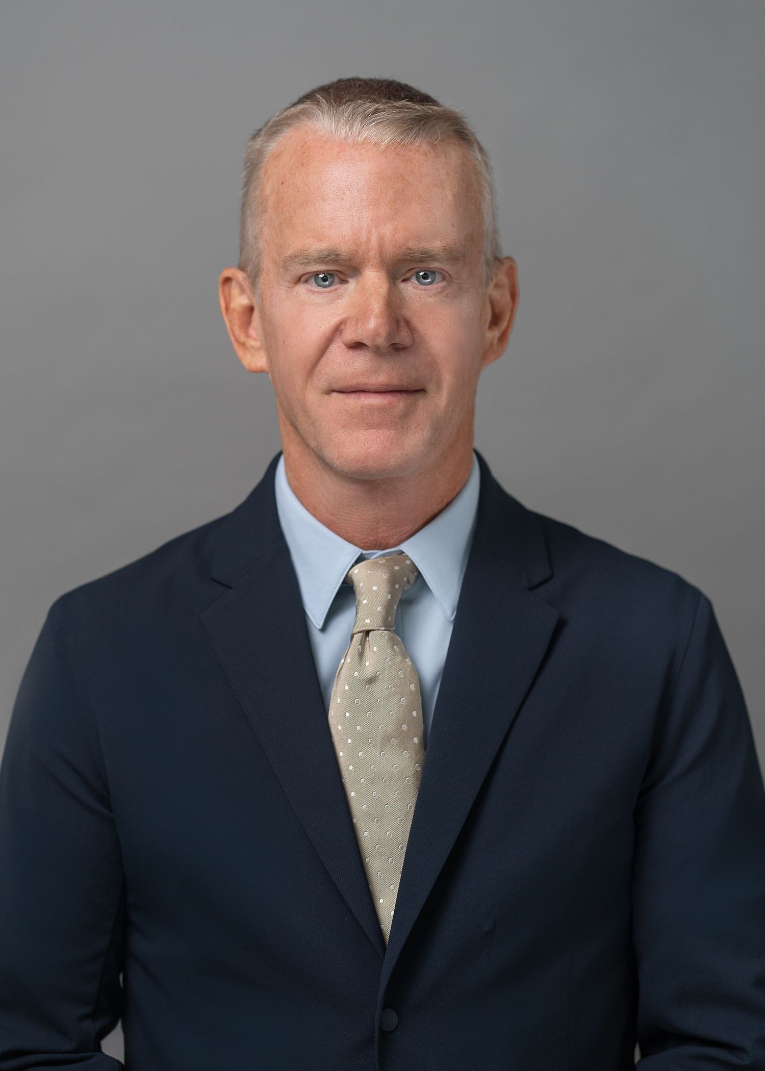 Professional headshot of businessman in navy suit and tie against grey background.