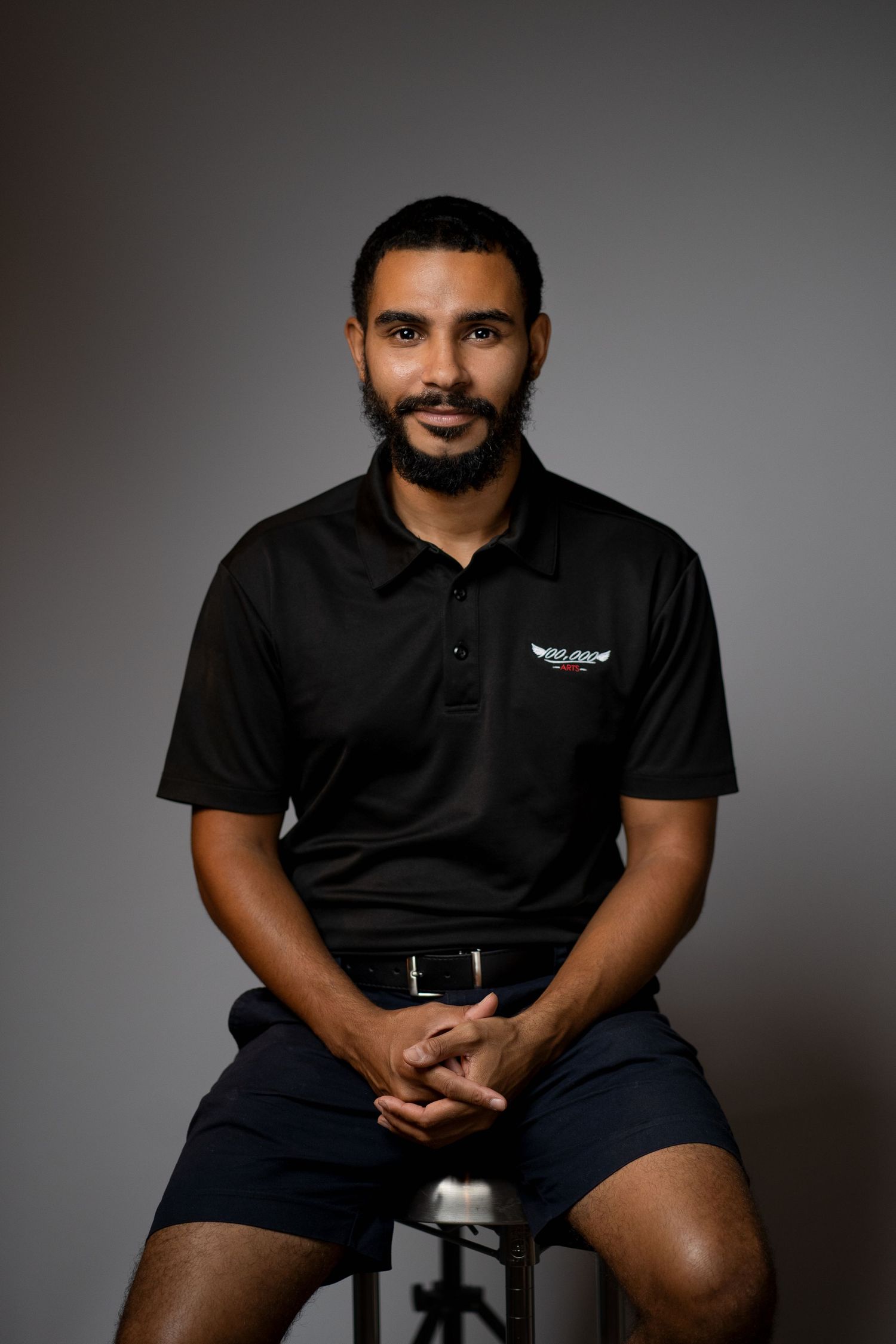 Professional seated portrait of person in black polo shirt against grey backdrop.