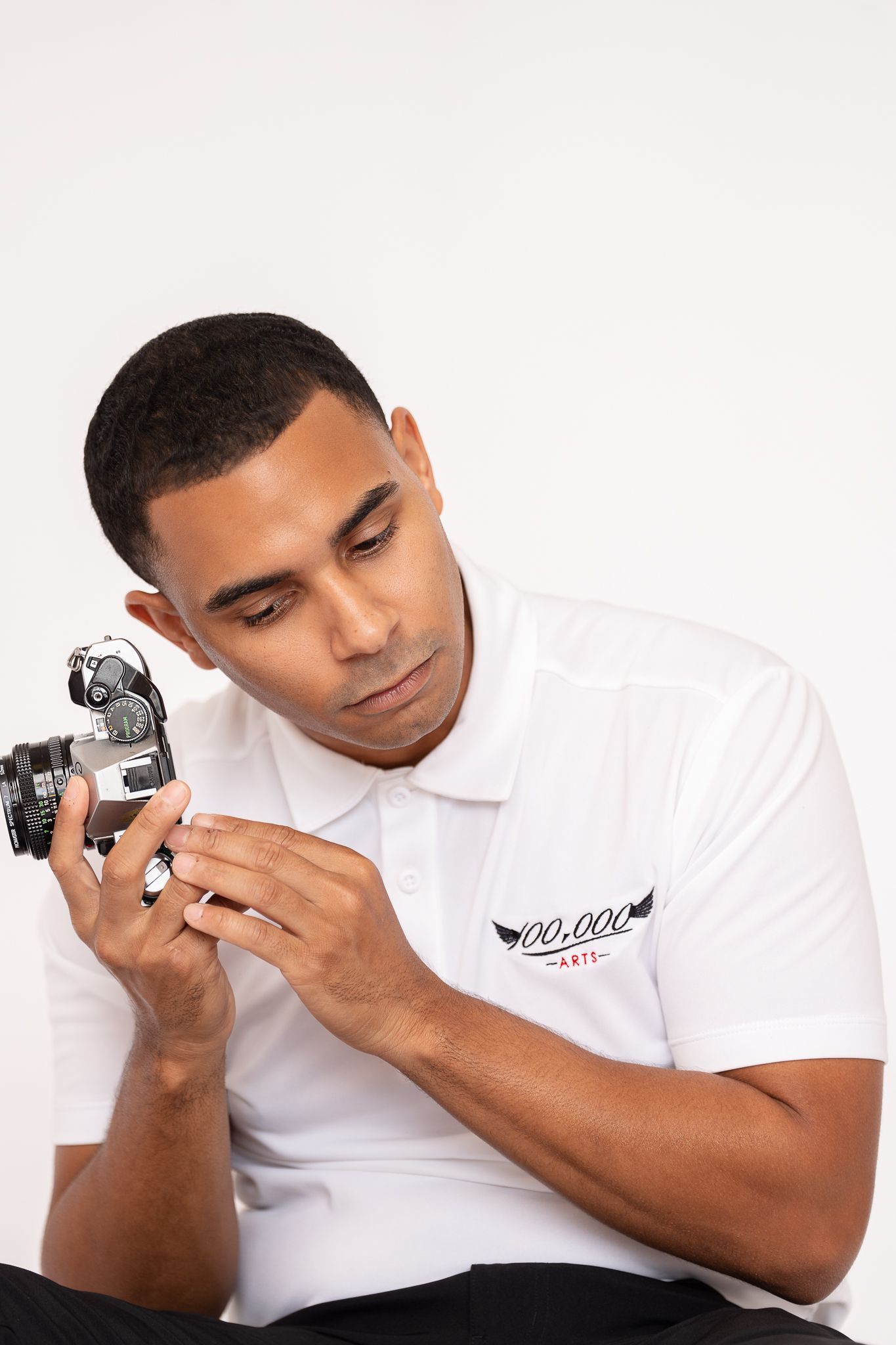 Person in white polo examining vintage camera while seated against white background.