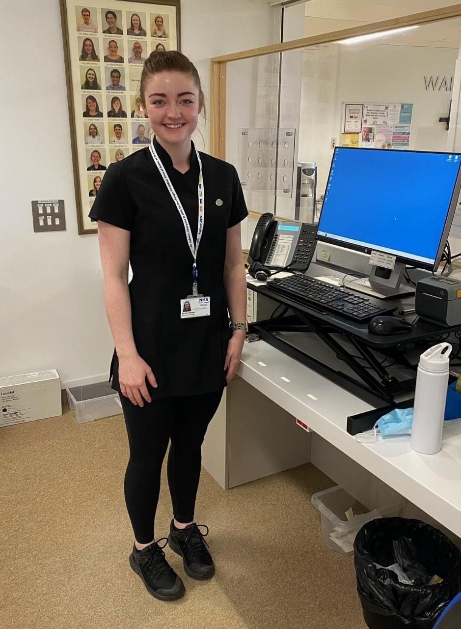 Staff member in black uniform stands at computer workstation in office setting.