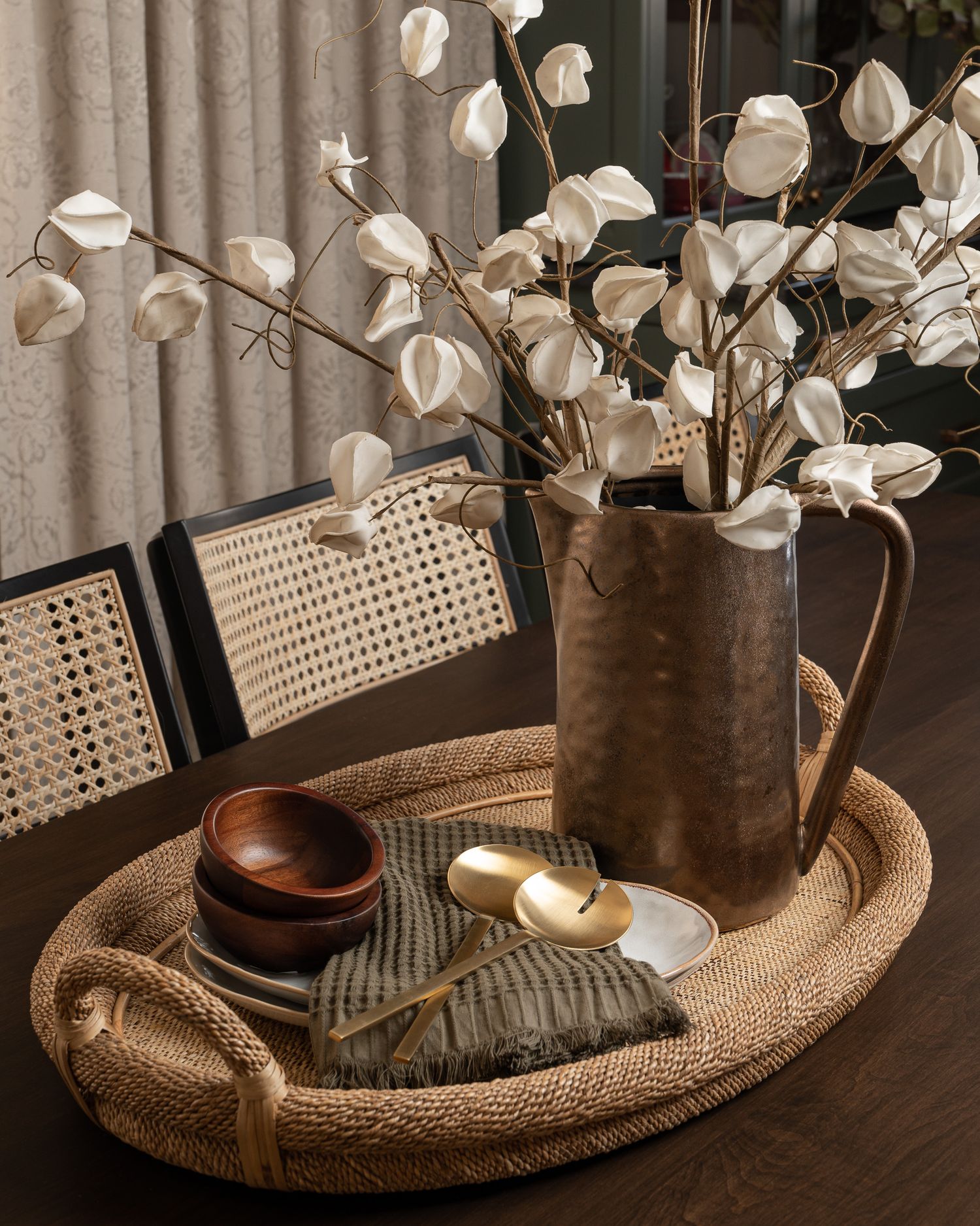 Rustic vignette featuring dried white flowers in brown pitcher on woven tray with wooden bowls.