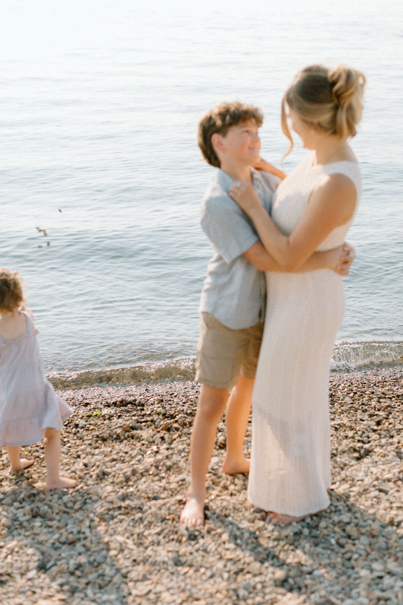 A mother and son share a tender embrace on a rocky beach shore while a young girl plays nearby by the calm sea.