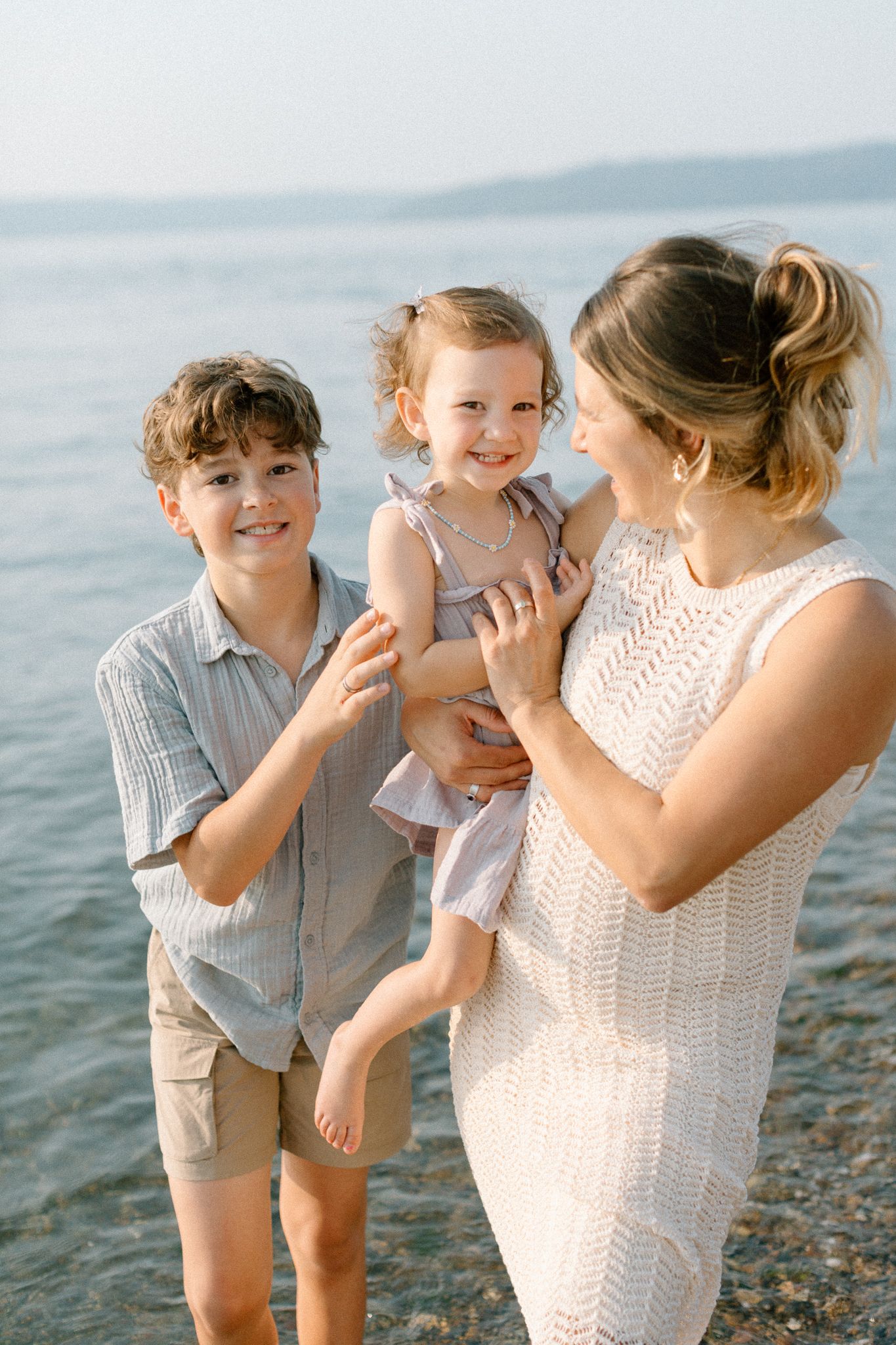 A mother holds her toddler girl while her young son smiles beside them on a sunny beach.