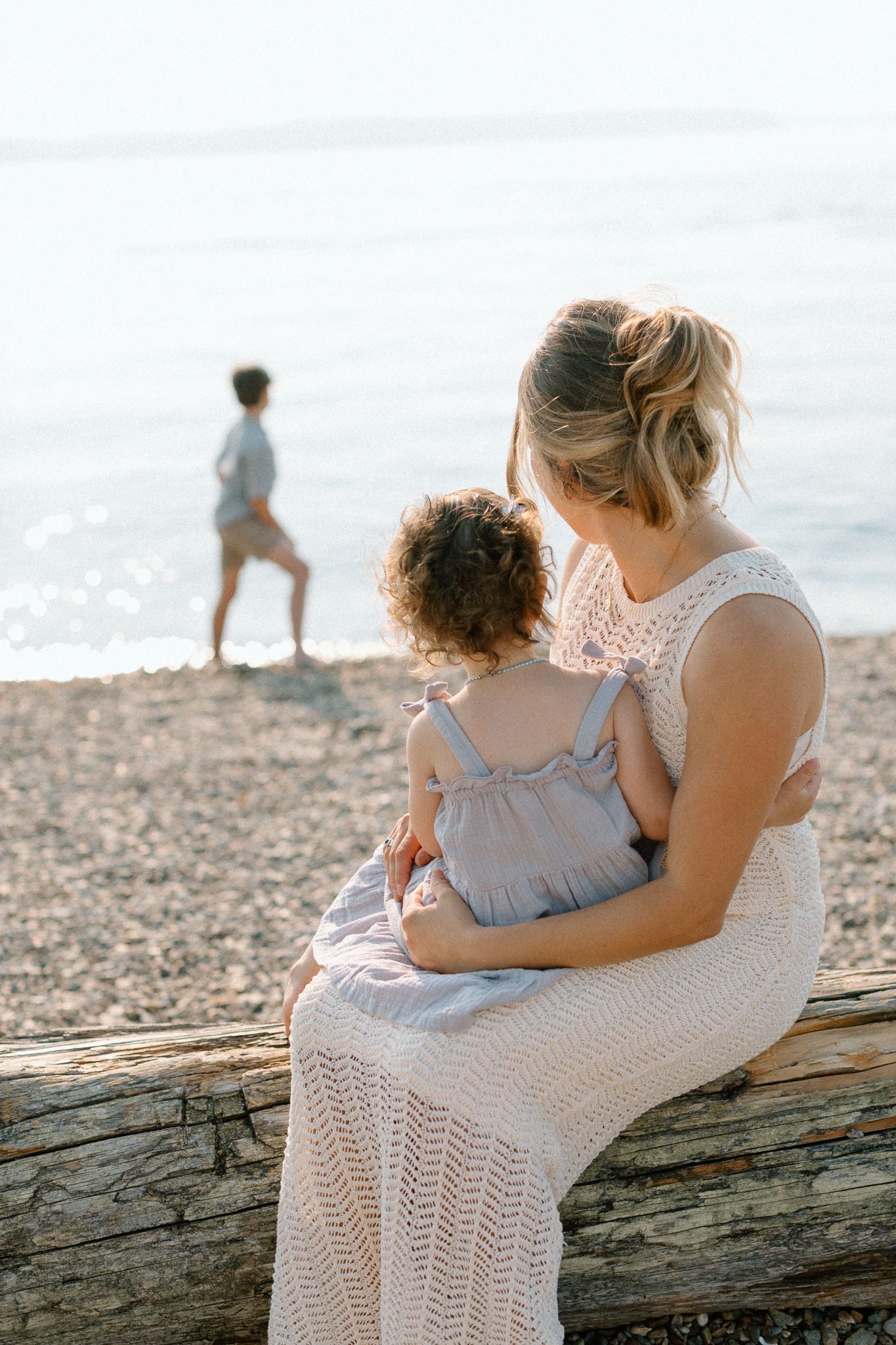 Mother in white lace dress holds toddler on driftwood log while child plays near ocean on pebble beach.