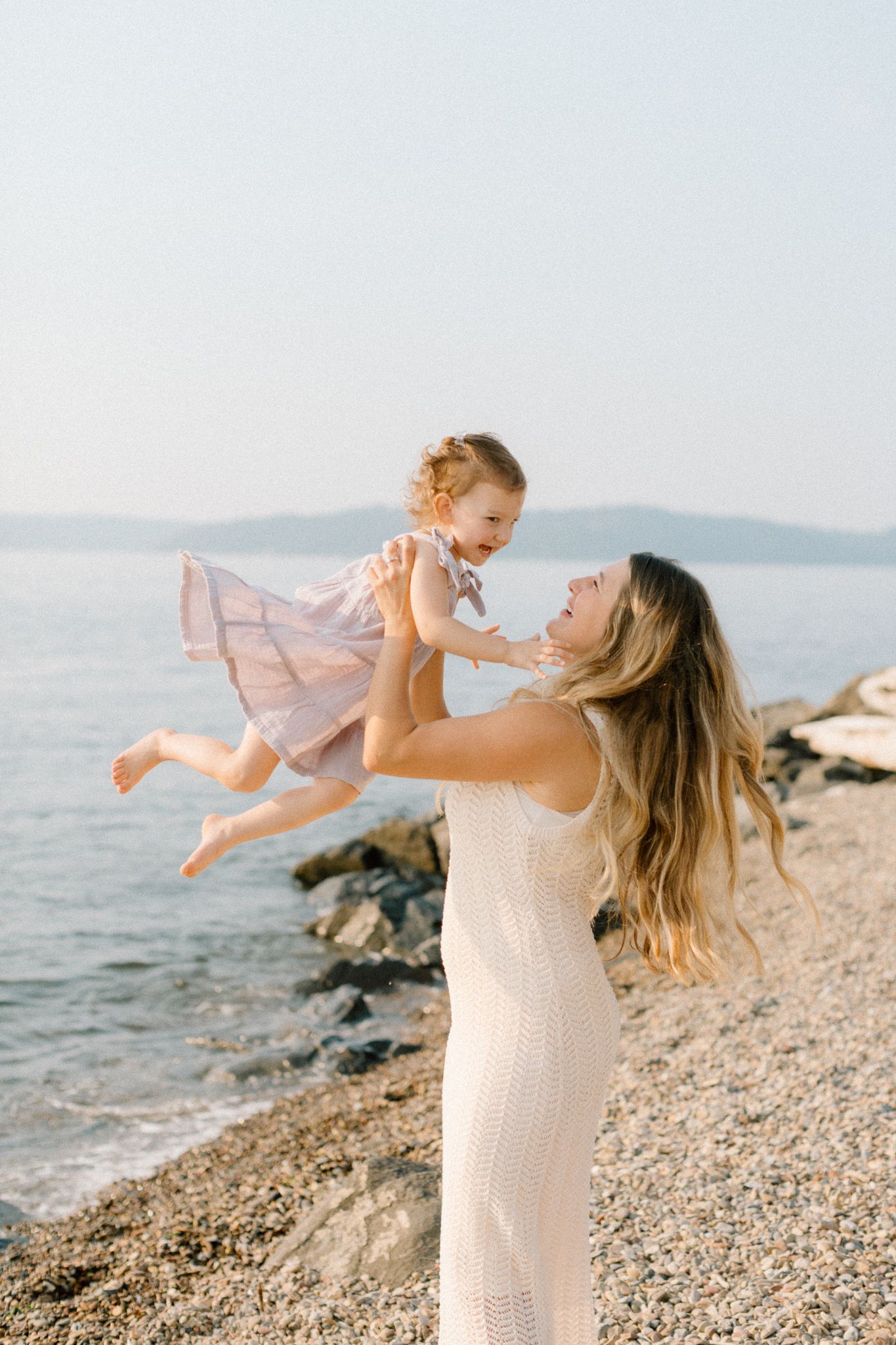 A mother in a white dress joyfully lifts her baby girl in a pink dress on a rocky beach by the sea.