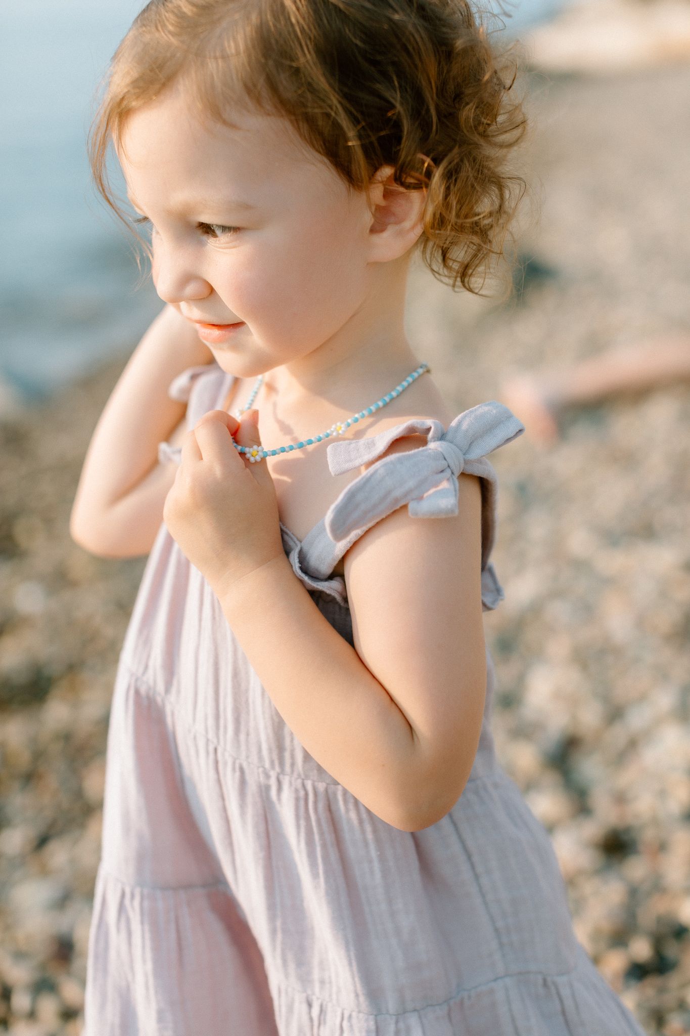 A toddler girl in a pink dress with bow straps and a beaded necklace smiles on a pebble beach.