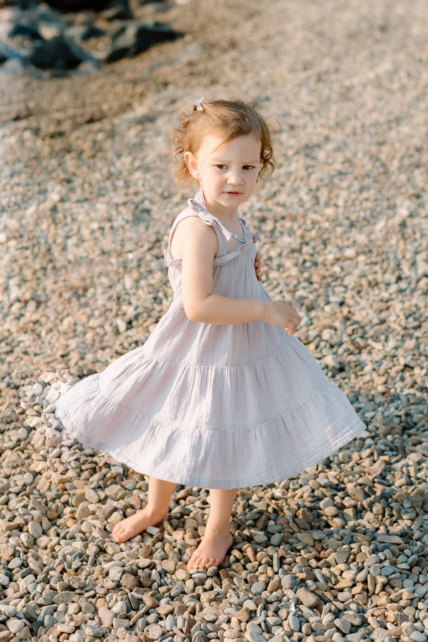 Adorable toddler girl in a light pink dress standing barefoot on a pebble beach, looking at the camera.