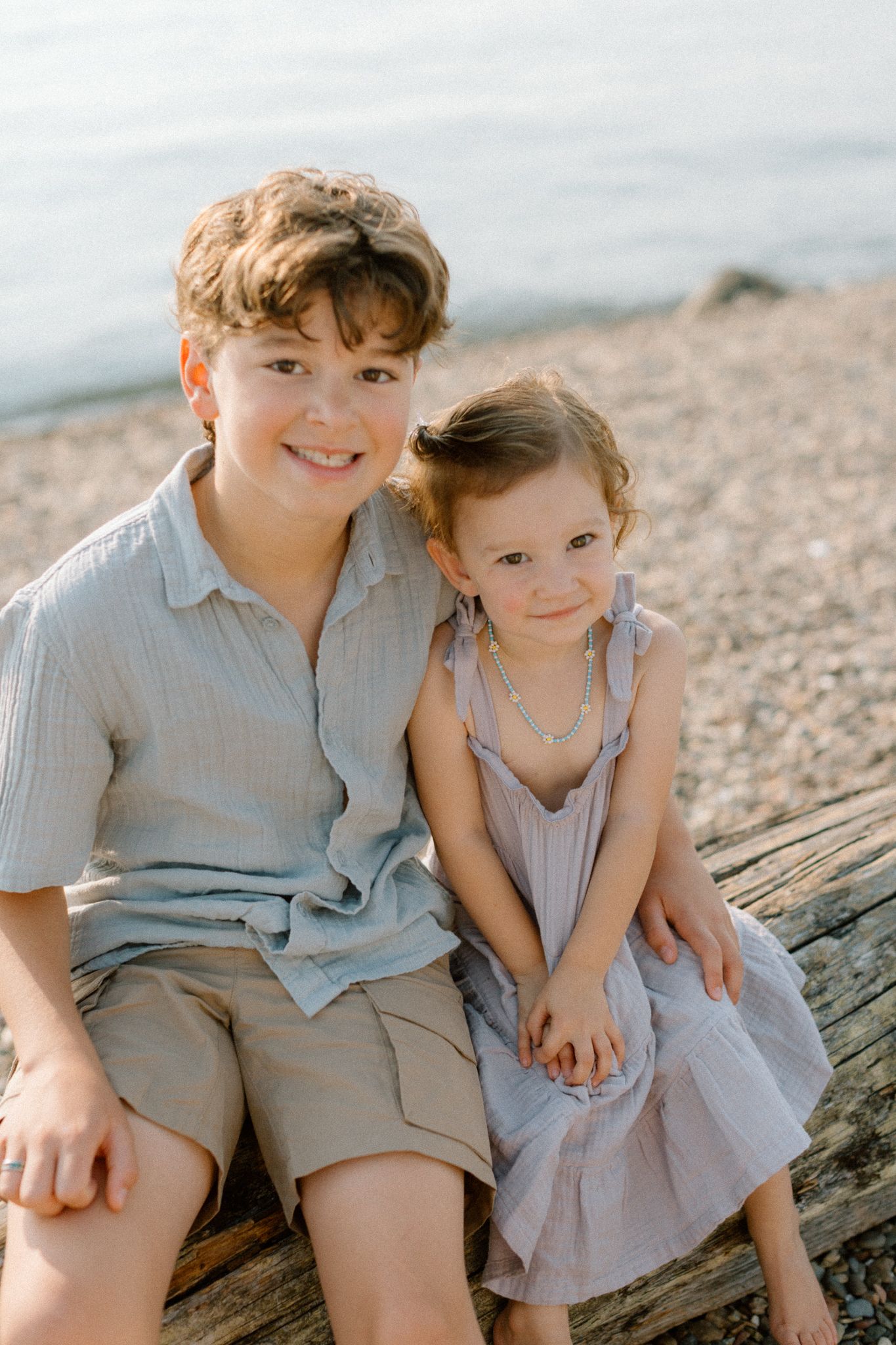 Two children sitting on driftwood at a rocky beach, older boy smiling with arm around younger girl in lavender dress.