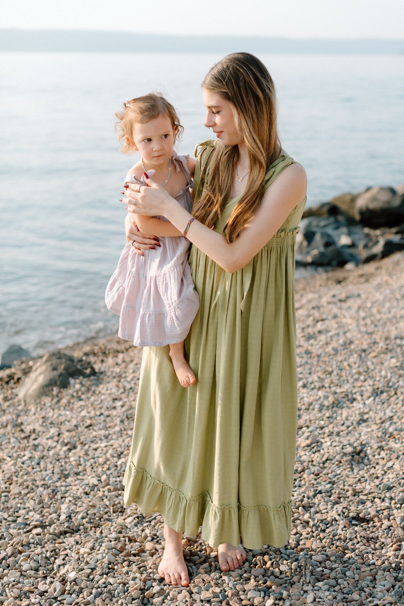 A young woman in a green maxi dress holds a toddler girl on a rocky beach near calm blue water.
