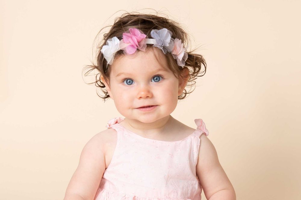 Baby in pink dress wearing floral headband against beige backdrop.