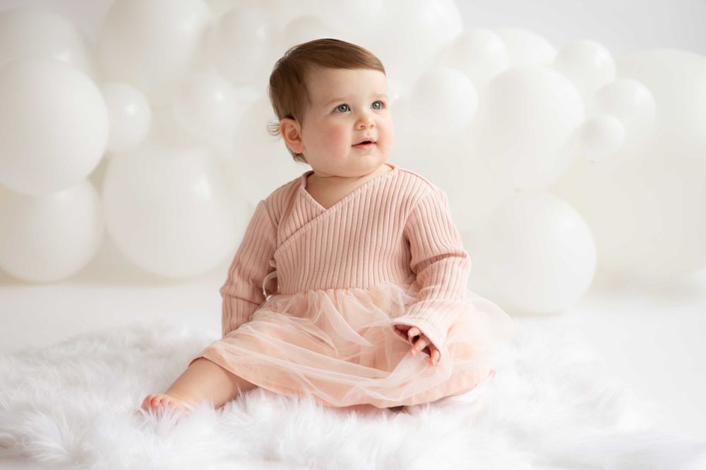 Baby in soft pink knitted dress sitting on white fluffy rug.