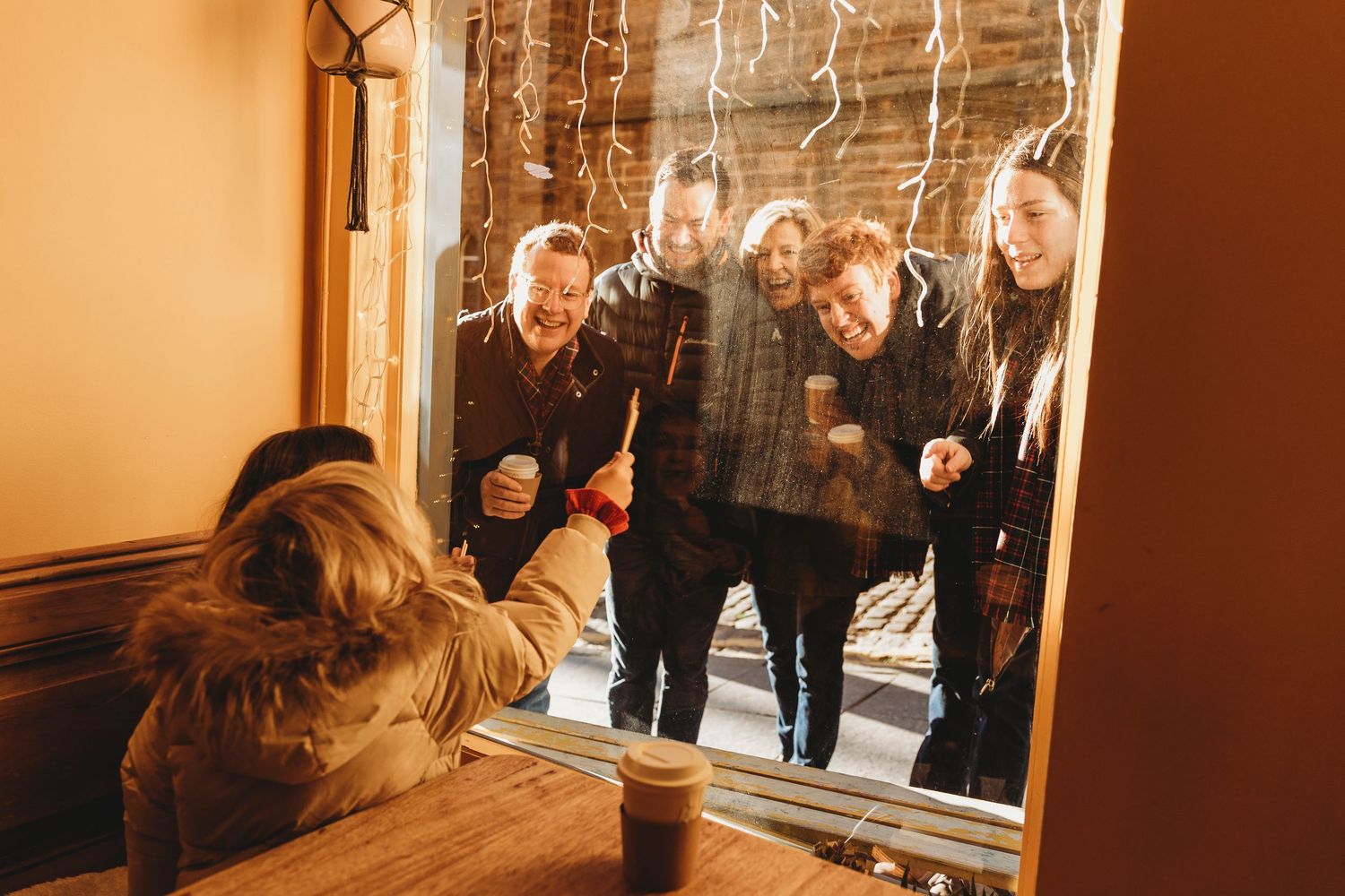 Friends peering through a window at someone inside with a coffee cup on a warm-toned afternoon.