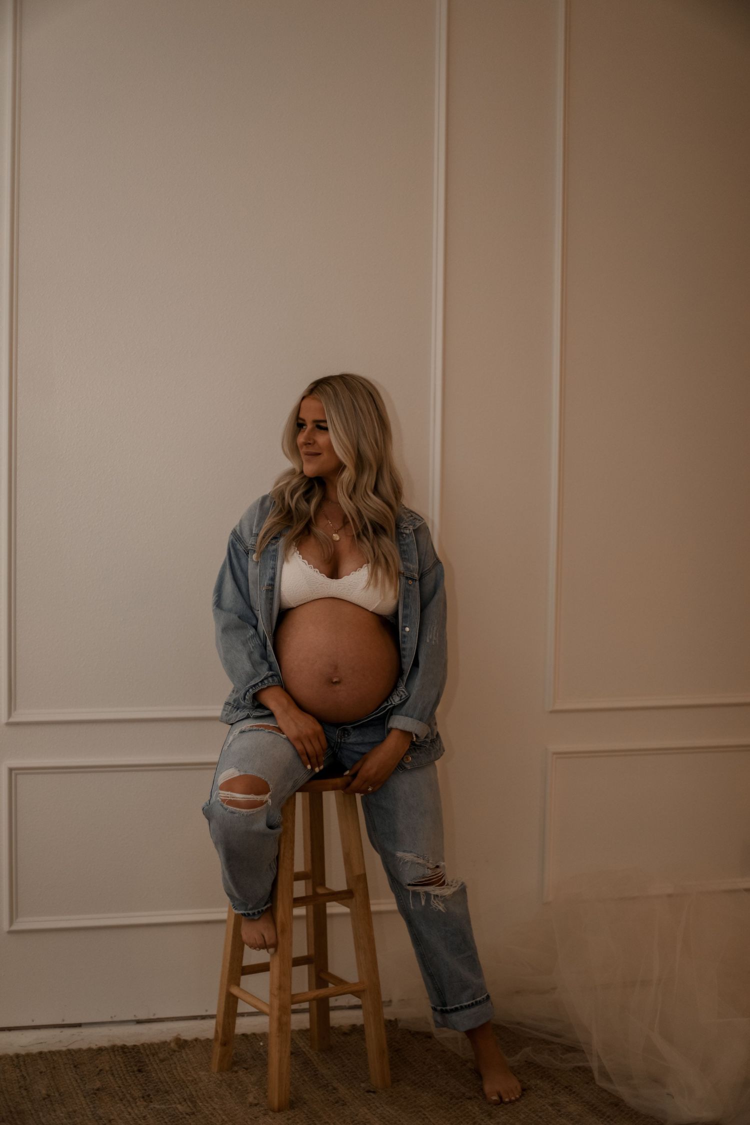 Casual maternity photo featuring distressed denim and wooden stool against neutral wall.