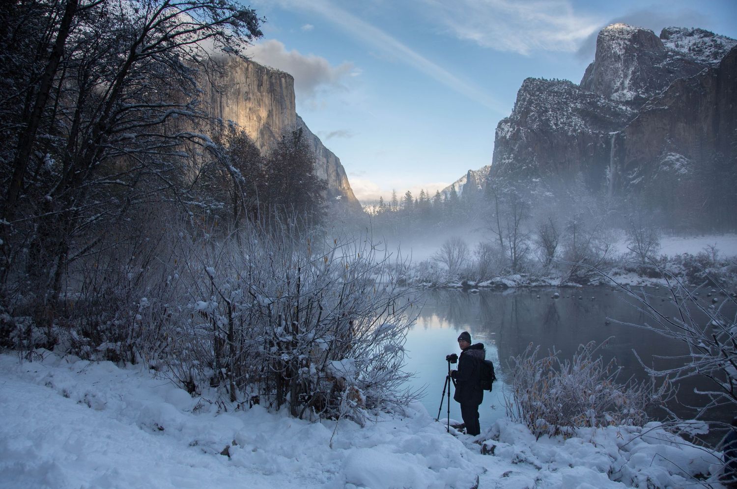 A silhouetted photographer captures the misty winter landscape of Yosemite Valley with snow-covered cliffs and reflective water.