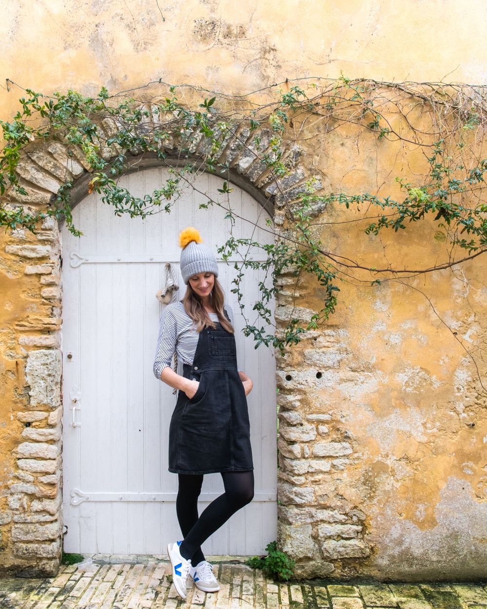 A person poses playfully against a weathered yellow stone wall with white door and climbing vines in a photo series.