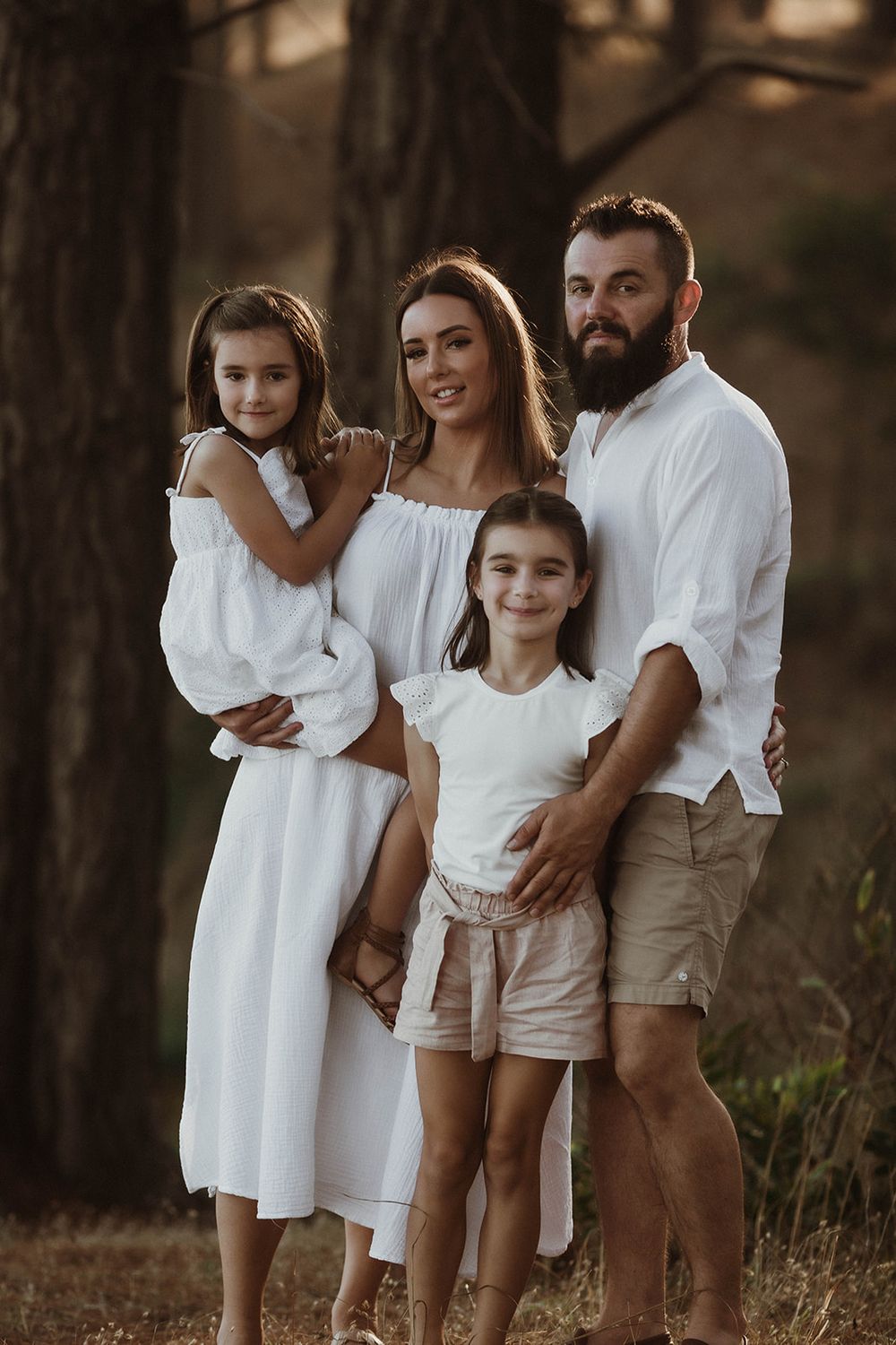 Family of four wearing coordinated white outfits poses among trees at sunset.