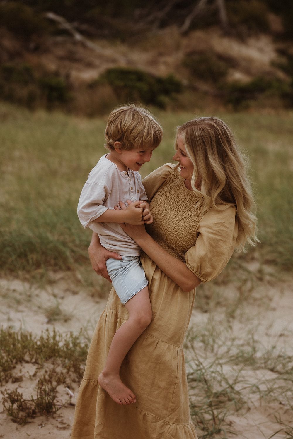 Mother in tan dress plays with young child on sandy ground.