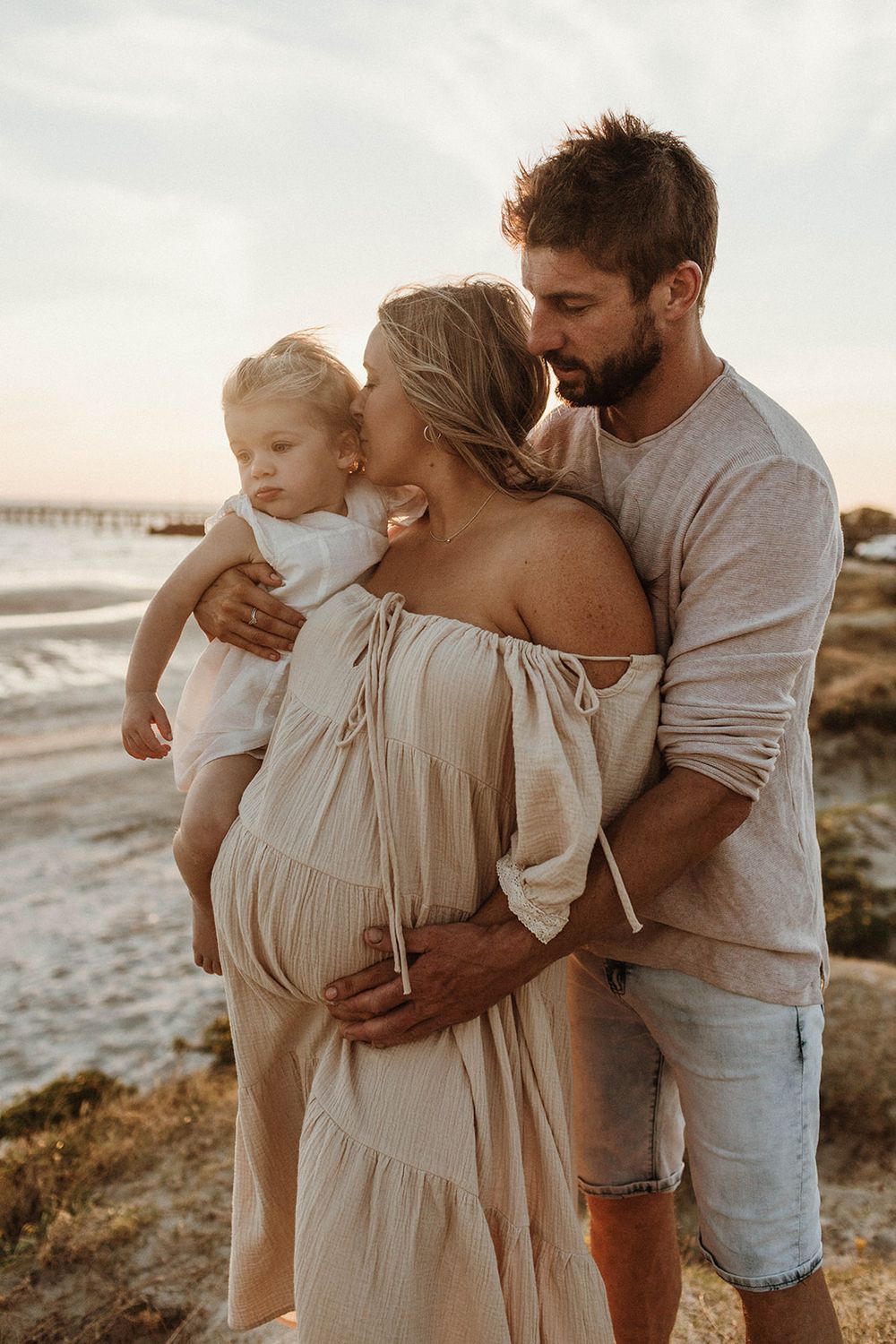 A tender family moment at sunset by the beach with a pregnant mother in a flowing dress.
