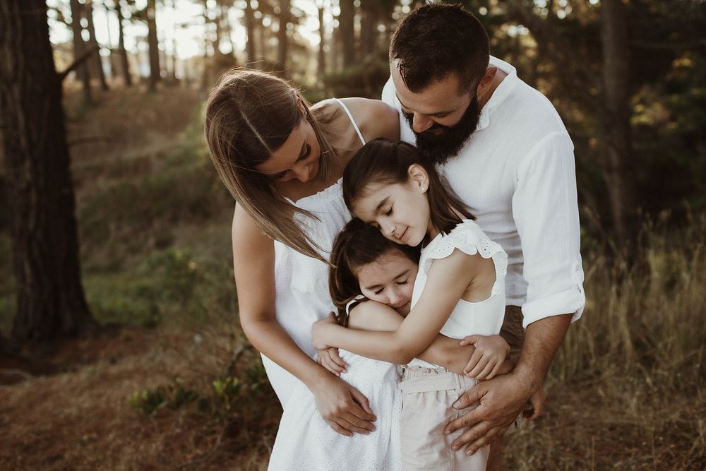 A family embraces in a warm group hug against a backdrop of pine trees at sunset.