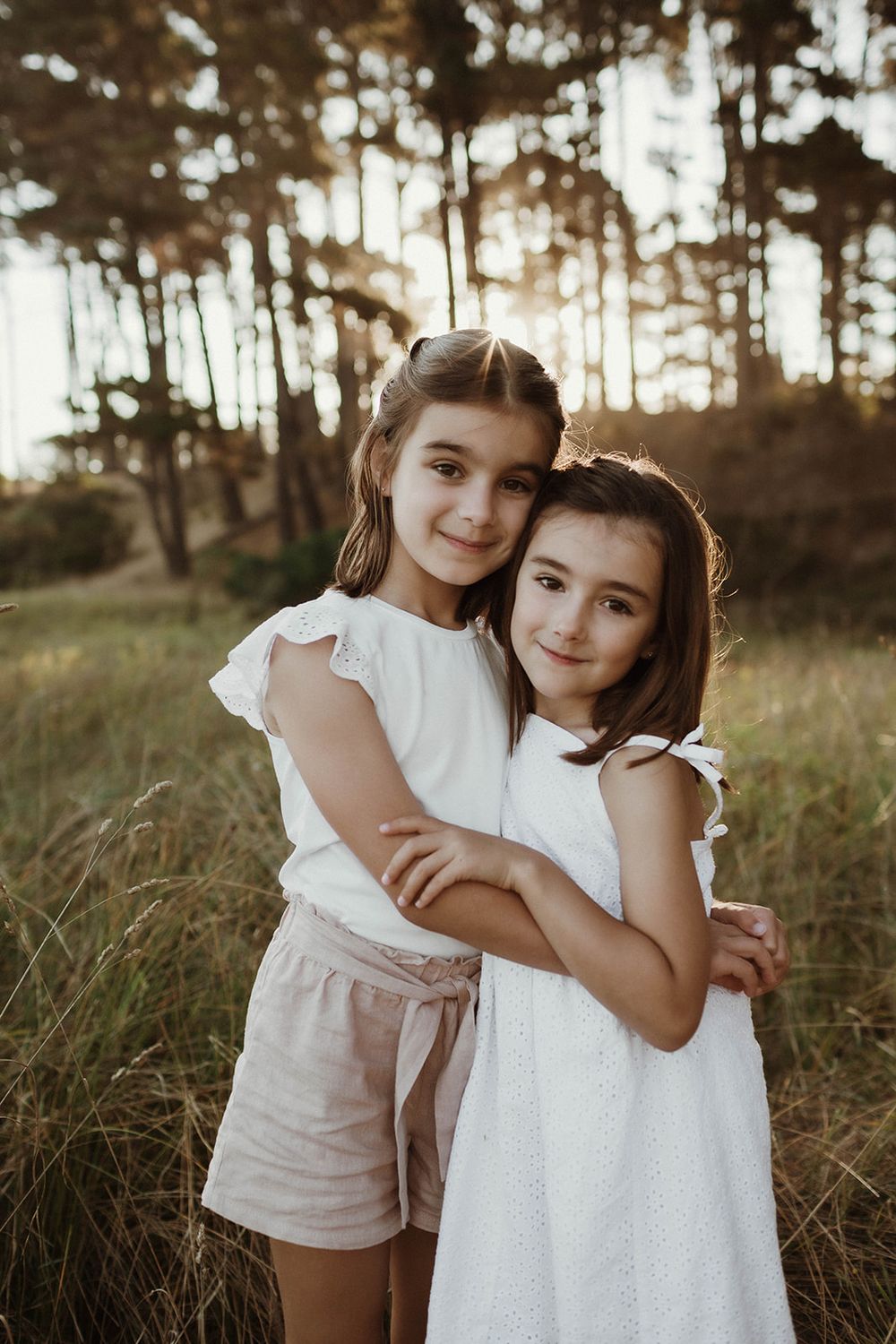 Two young people in white outfits pose together in a sunlit field with pine trees.