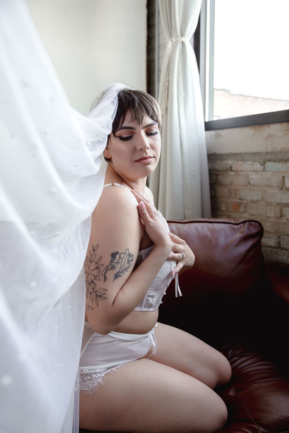 Natural light boudoir photography in loft space with exposed brick walls and sheer curtains.