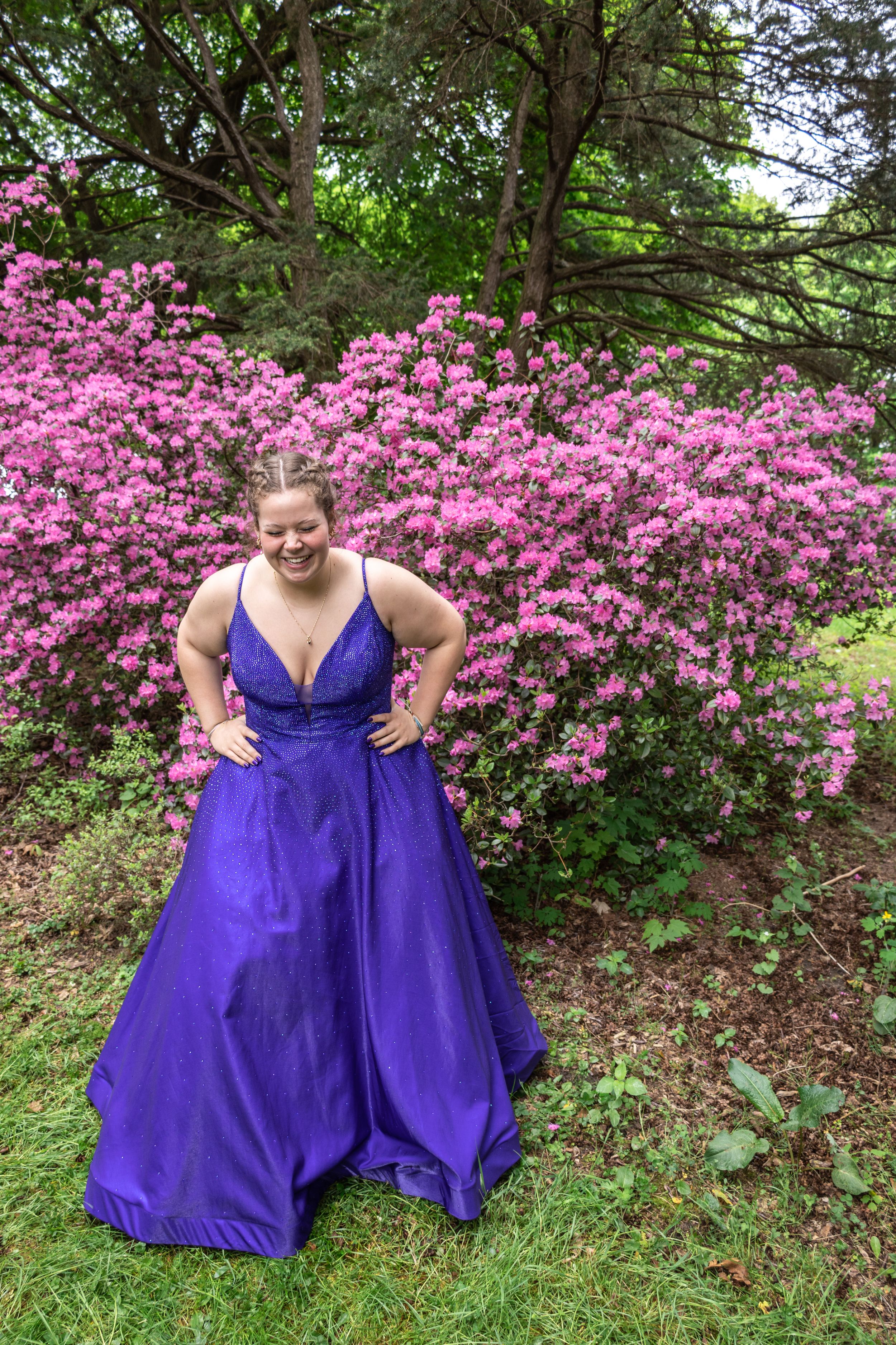 A person in a flowing purple gown poses confidently against pink flowering bushes.