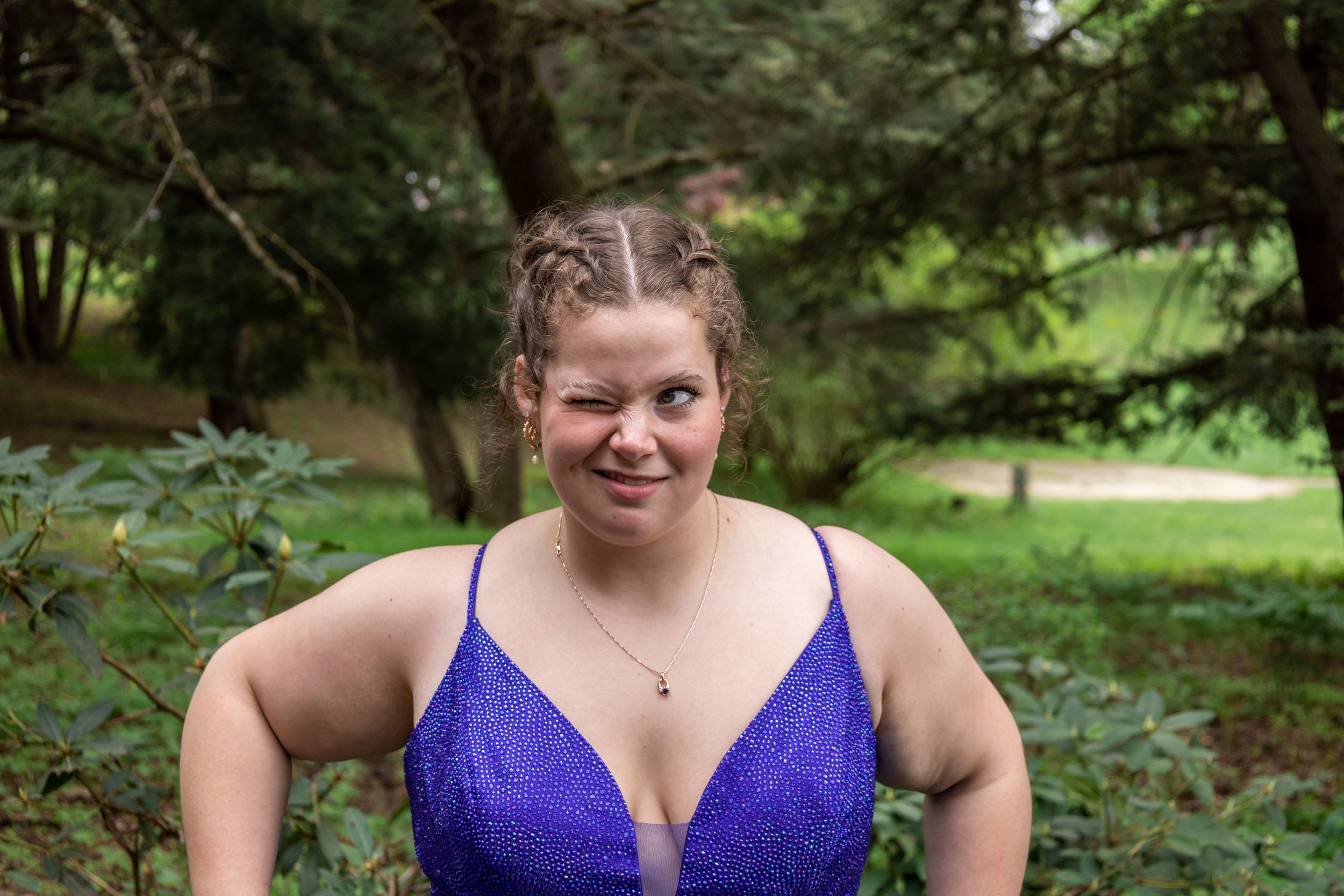 A person in a sparkling purple formal dress poses in a lush garden setting.