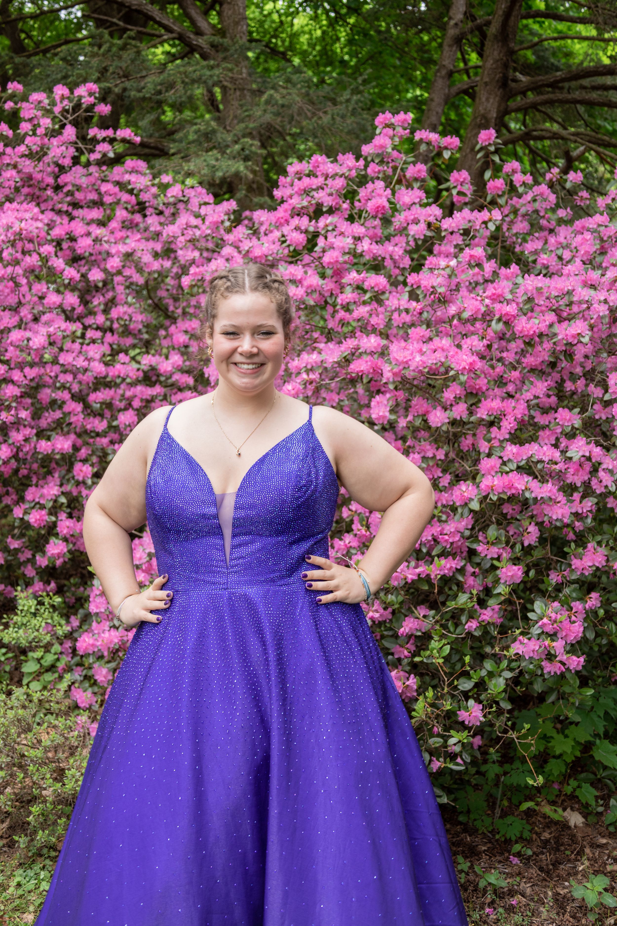 Purple formal gown photographed against blooming pink azalea bushes in a garden setting.