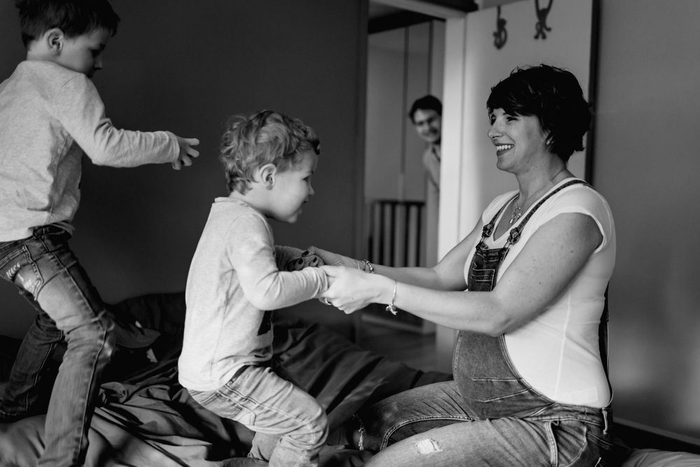 maman enceinte joue avec ses ainés sur le lit dans la chambre parentale sous le regard attendri du papa en séance photo