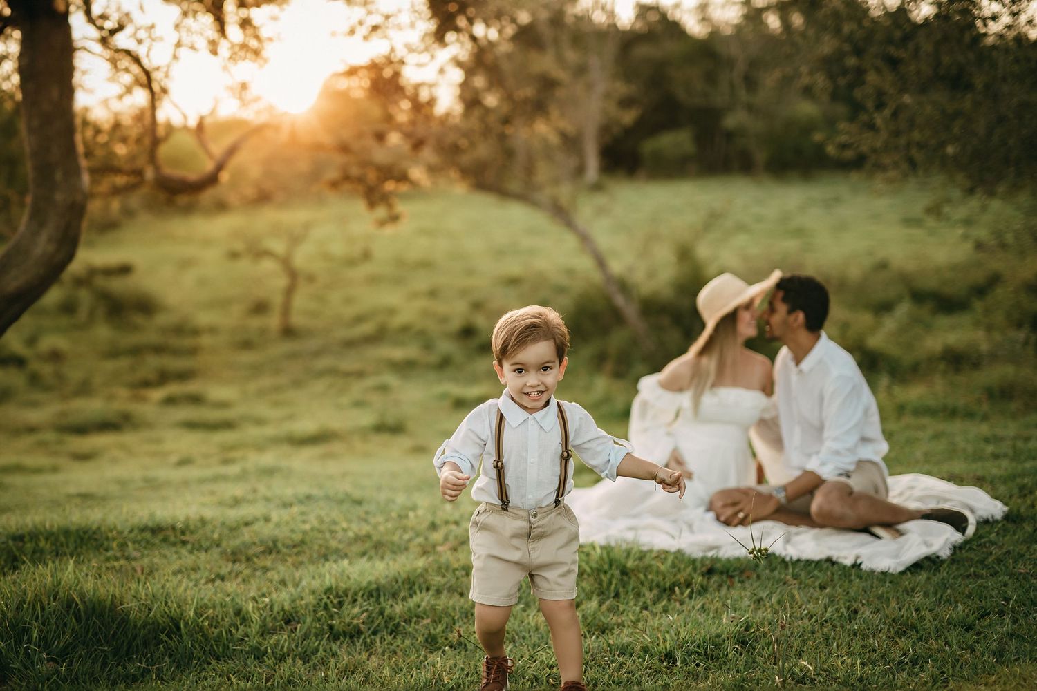 Family enjoys sunset picnic on grassy hill while toddler plays in foreground.