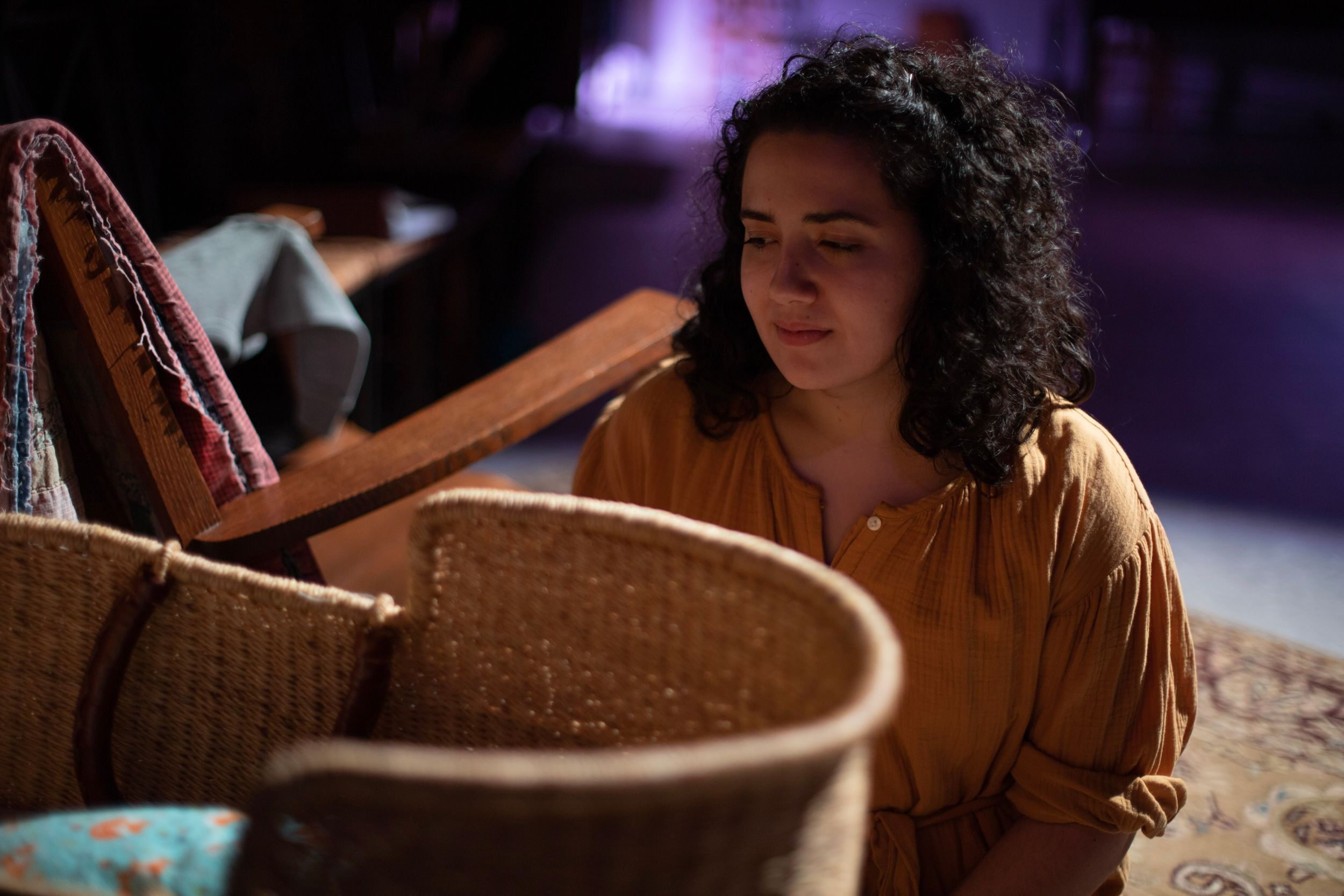 A performer in an orange dress examines a woven bassinet in atmospheric backlighting. From Mary's Wondrous Body in 2022.