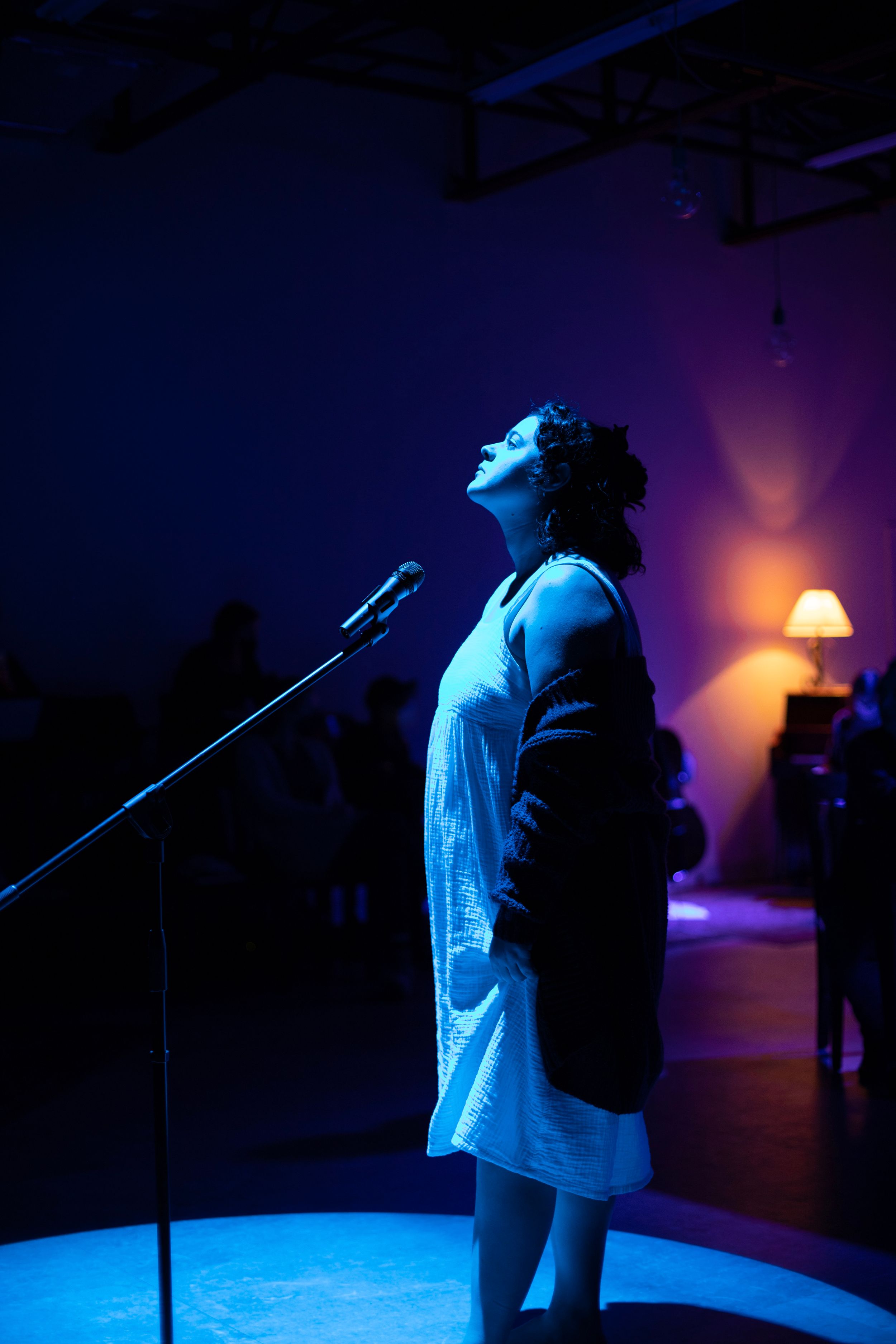 A performer stands at a microphone bathed in moody blue and purple stage lighting. Production photo from Mary's Wondrous Body in 2022.