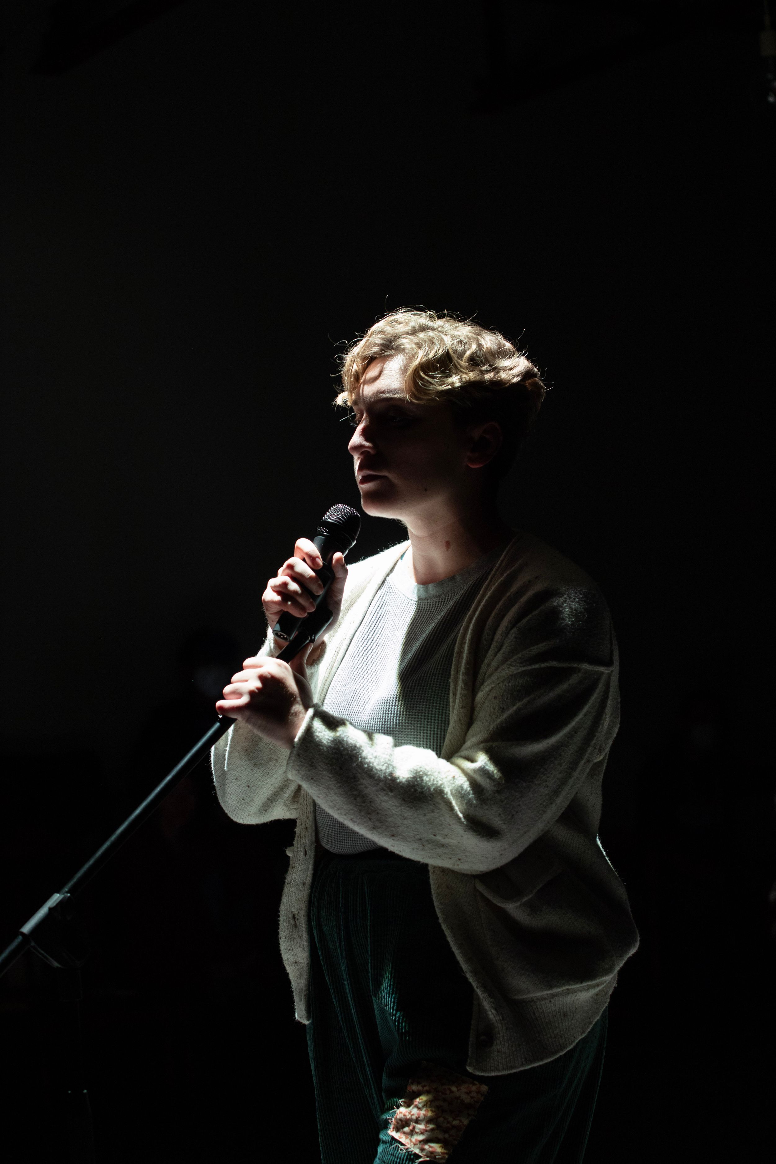 A performer in a cardigan holds a microphone while singing on a dramatically lit stage. Mary's Wondrous Body in 2022 at Elision Playhouse.
