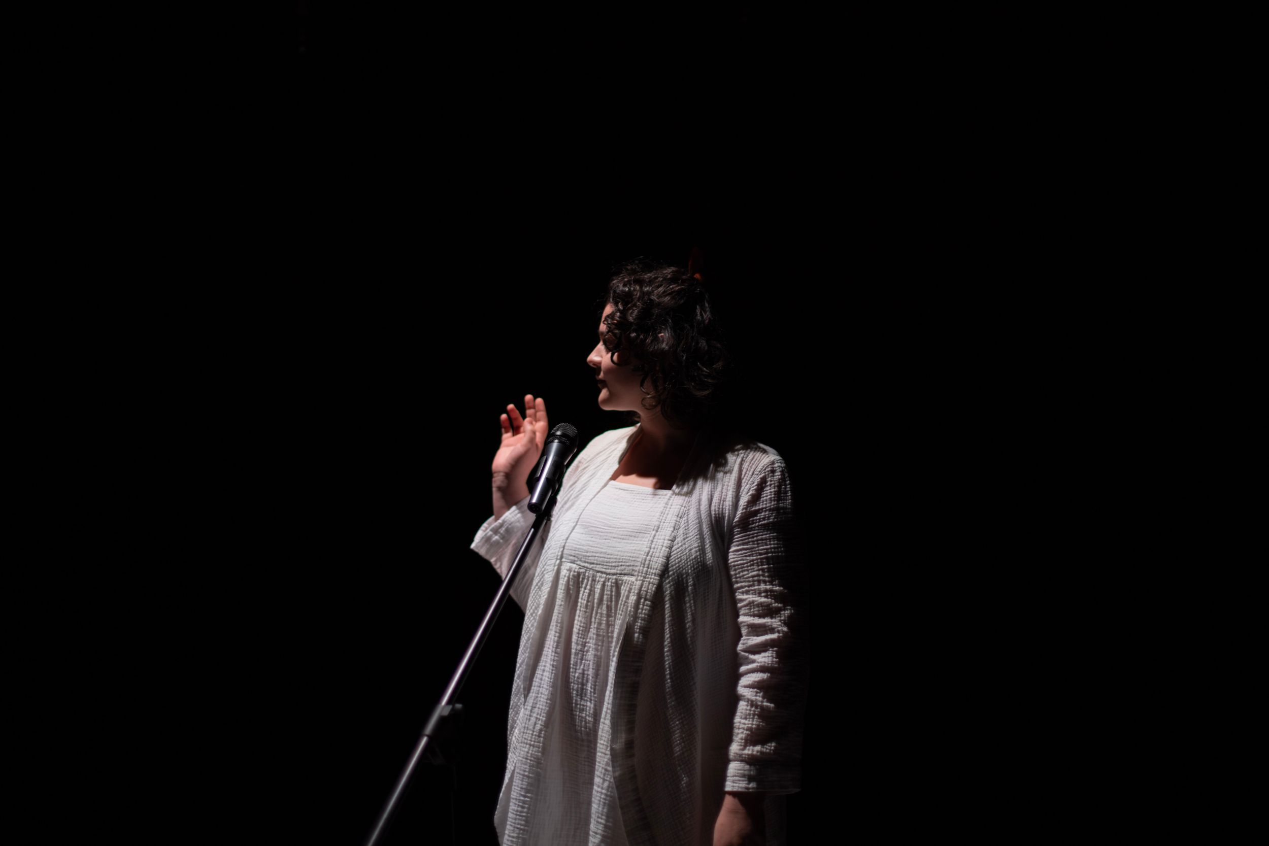 A performer in a white dress sings on stage against a black background. From Mary's Wondrous Body in 2022.