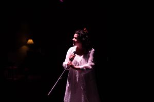 A solo performer in a light-colored dress sings passionately on stage in dramatic lighting. From Mary's Wondrous Body in 2022.