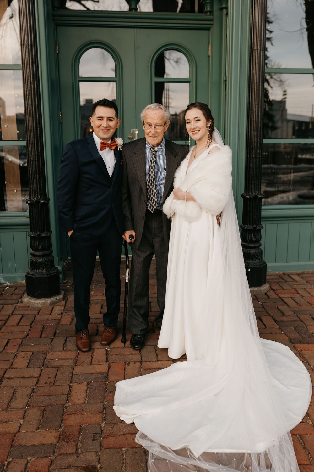 Bride and groom pose with elderly gentleman outside green historic building on brick walkway.