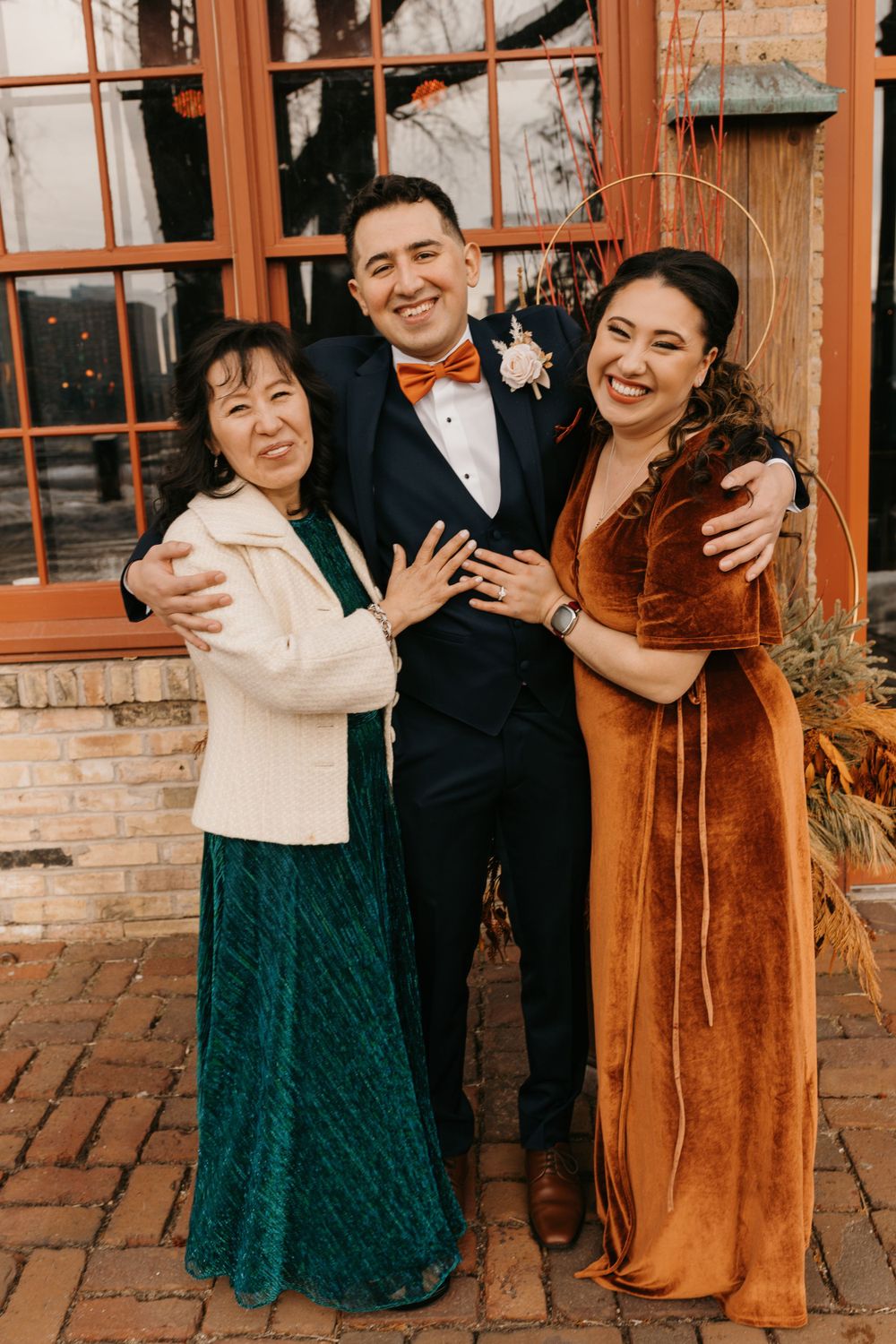 Groom poses with two women in rust velvet and teal dresses at venue.