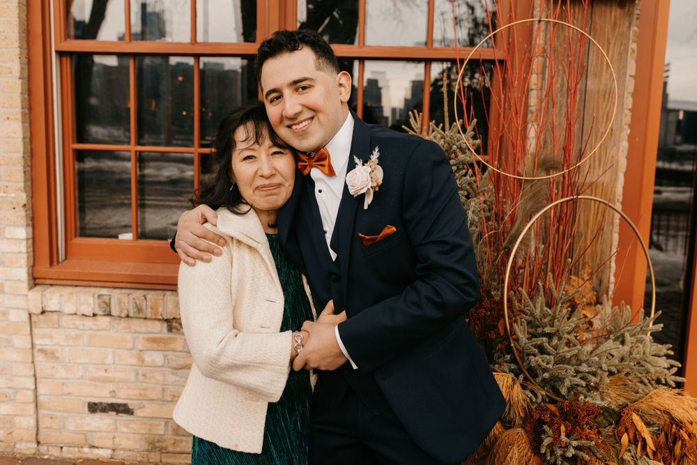 Groom in navy tuxedo embraces older woman in white jacket outside brick venue.